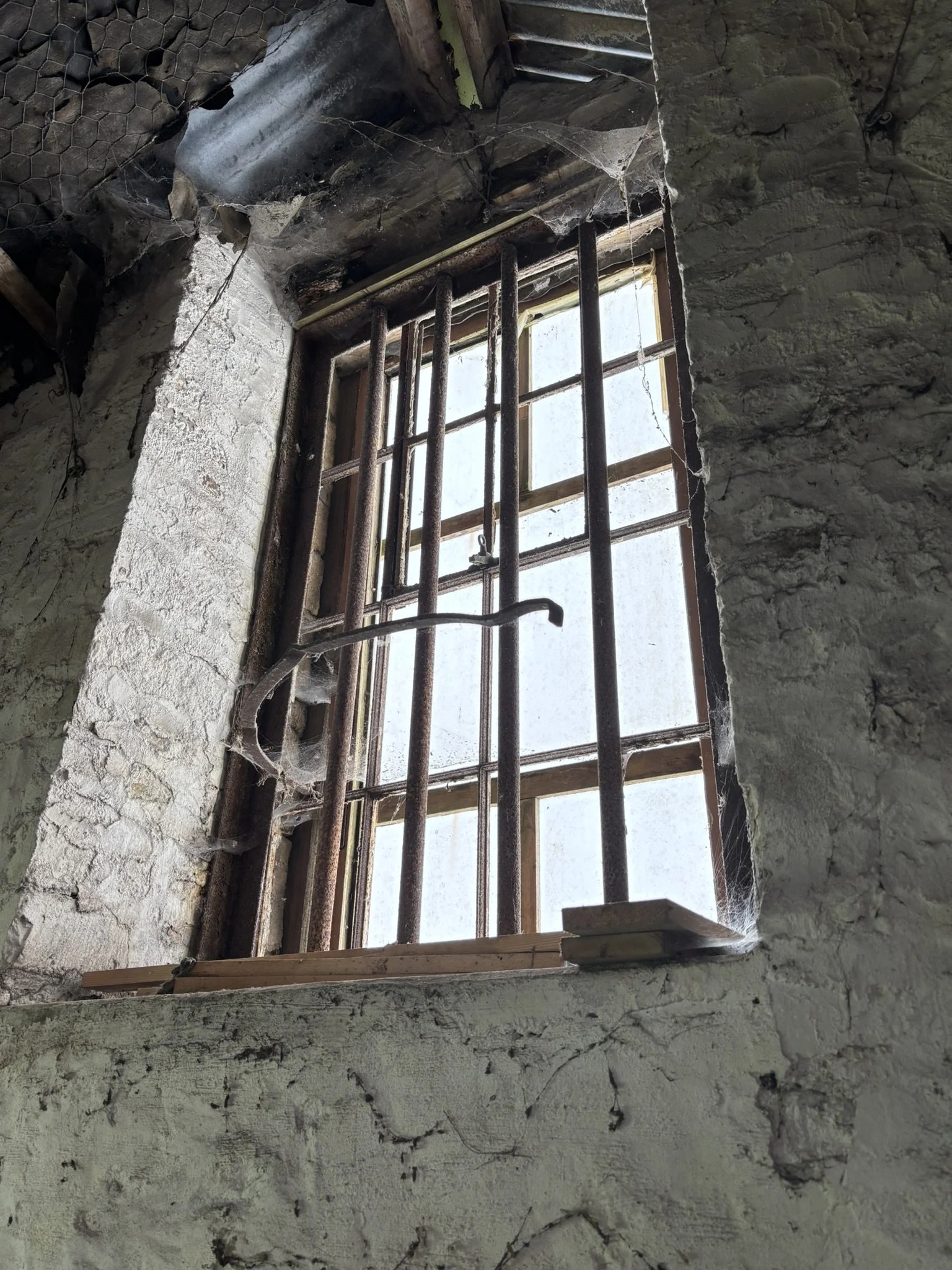 A small barred window in a dilapidated stone wall with cracks and peeling paint, cobwebs, and some wooden trim around the window, viewed from inside a run-down building.