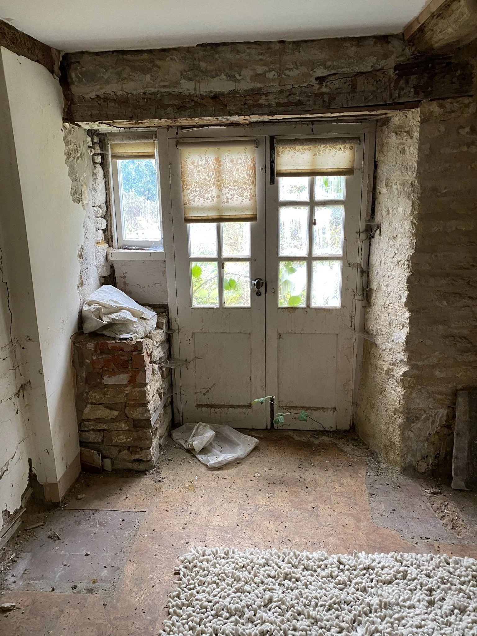 A room with partially exposed brick and stone walls, a wooden beam, and a dirt-covered floor. There is a window with two sliding panels, each covered with a beige, floral-patterned blind. Some debris and a pile of rocks are visible near the window, and a curtain of small white objects is on the floor in the foreground.