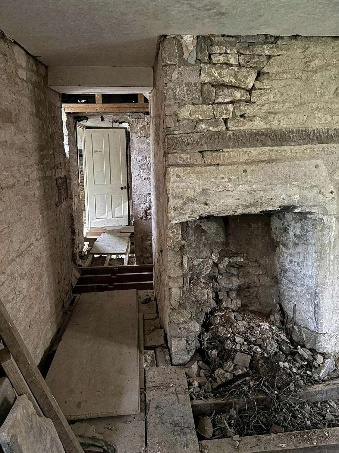 Photo of the inside of a dilapidated building. A brick walkway and fireplace with broken and removed floorboards. Looking to a white door at the exit.