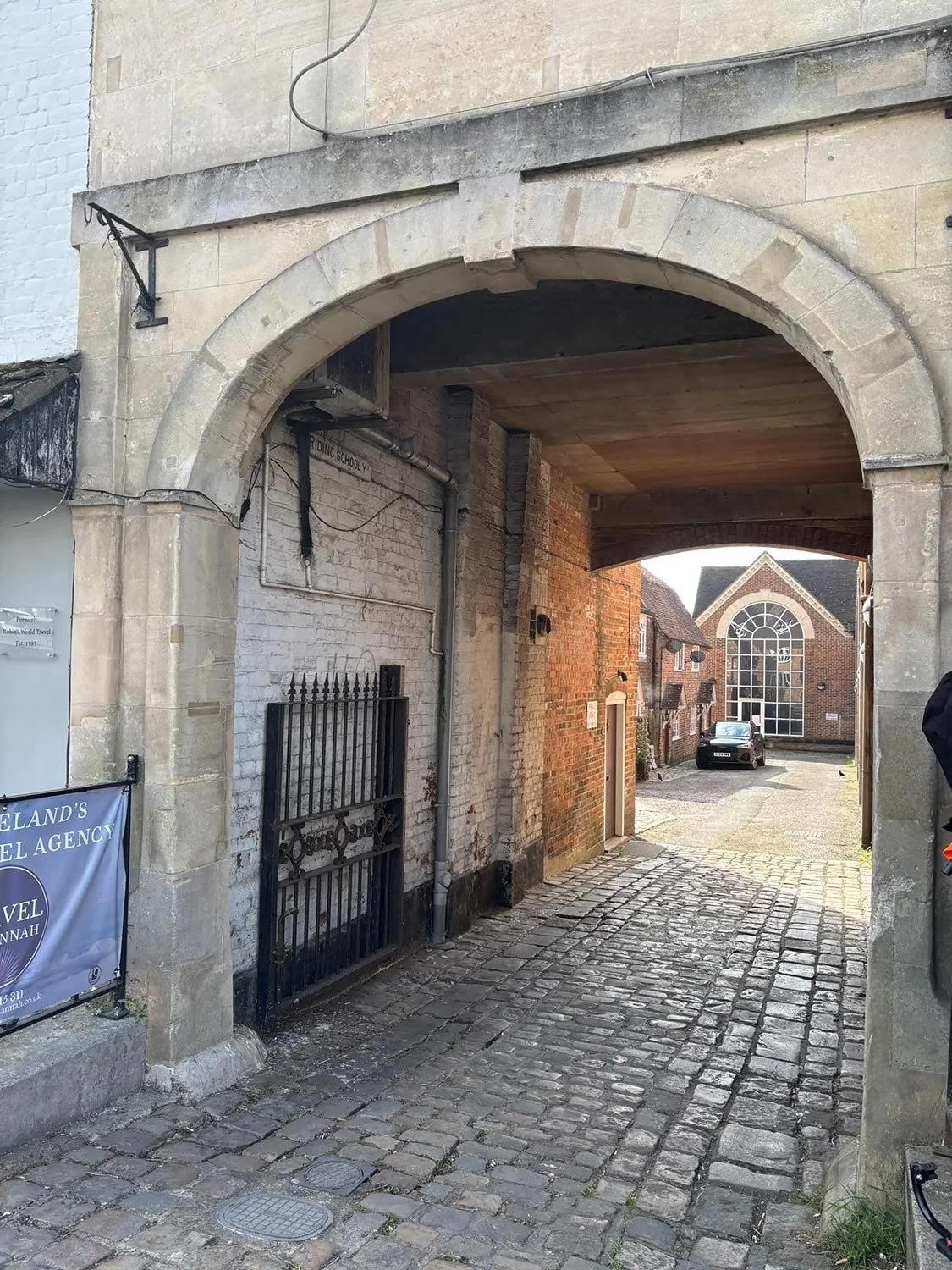A cobblestone alleyway framed by an old stone arch, leading to a small square with brick buildings in the background.