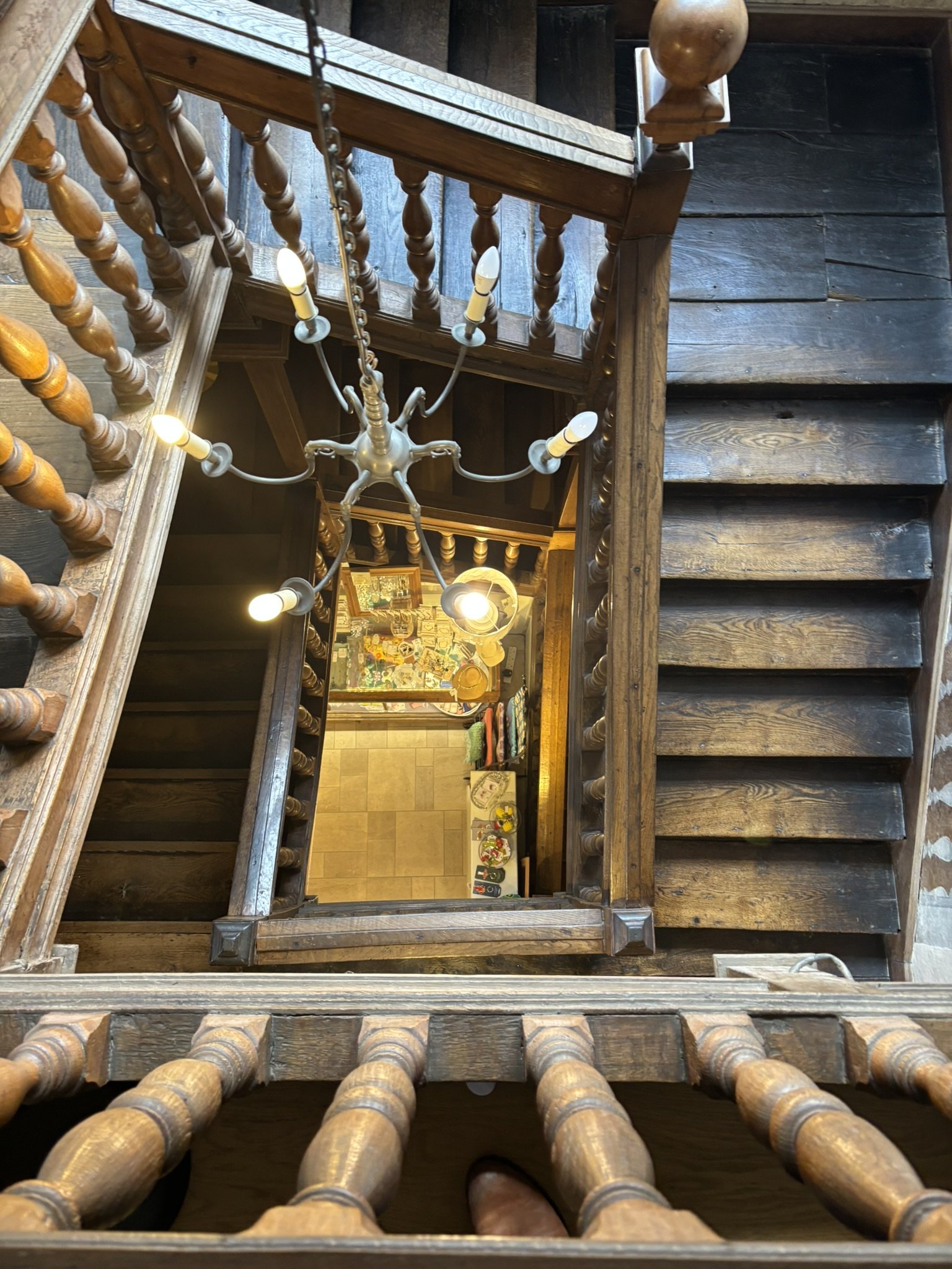 Top-down view of a wooden staircase with turned spindles, leading to a landing decorated with colorful items and memorabilia, illuminated by a chandelier.