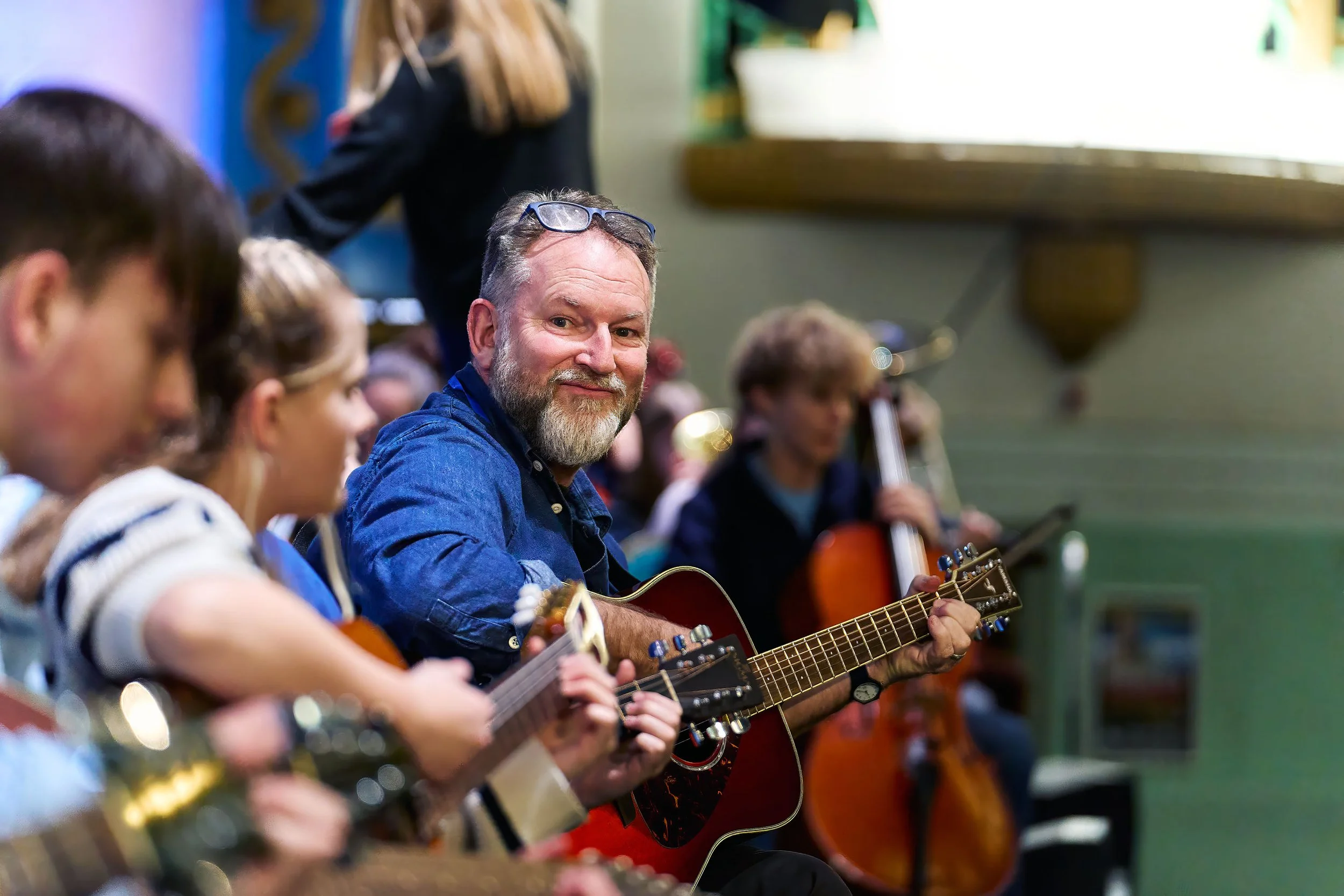 People sitting in a row playing acoustic guitars, with a man wearing glasses resting on his head, in a room with green walls and a window.