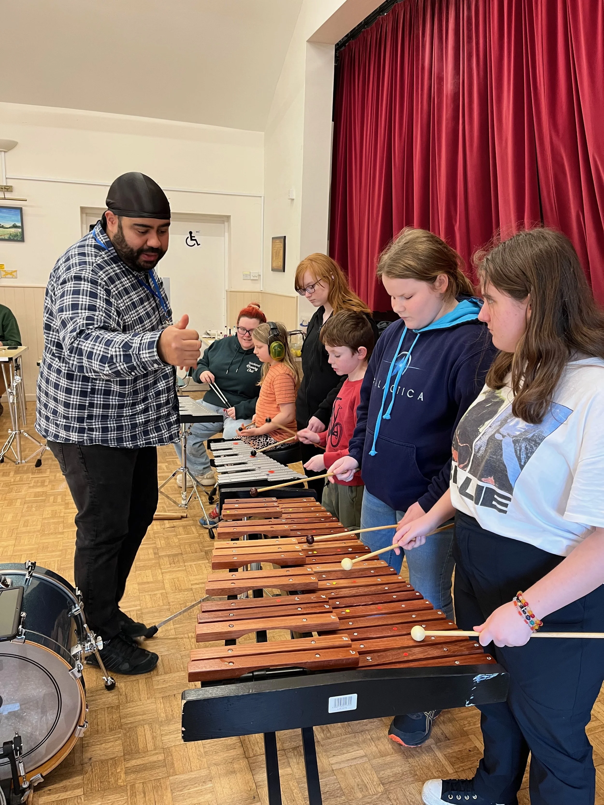 A group of children playing xylophones and percussion instruments in a room with red curtains and a man guiding them.