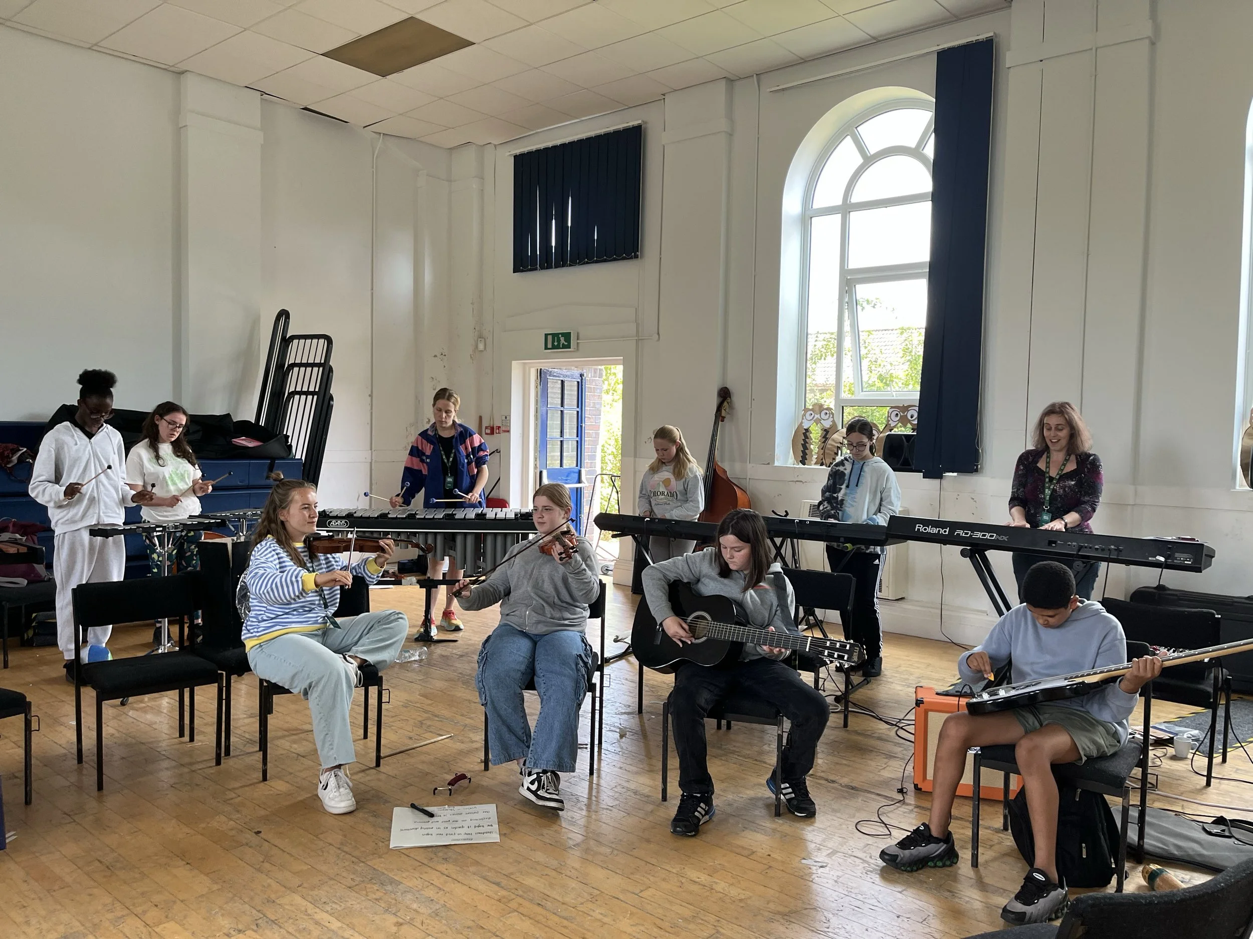 A group of children and a woman are in a music class in a bright, spacious room with large arched windows, some holding musical instruments, and a music teacher guiding them.