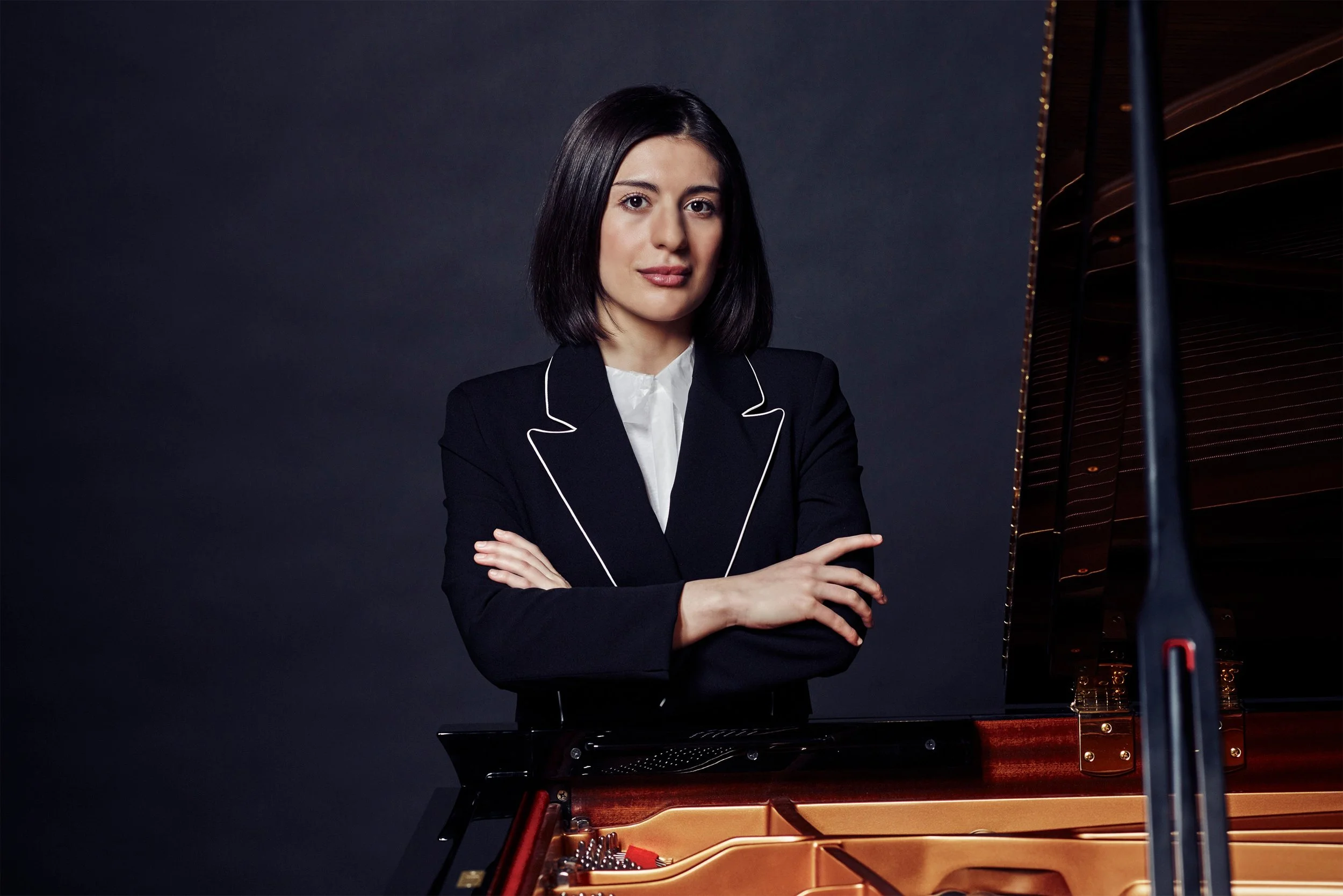 A woman with dark hair dressed in a black blazer and white blouse standing beside a grand piano, with her arms crossed, against a plain dark background.