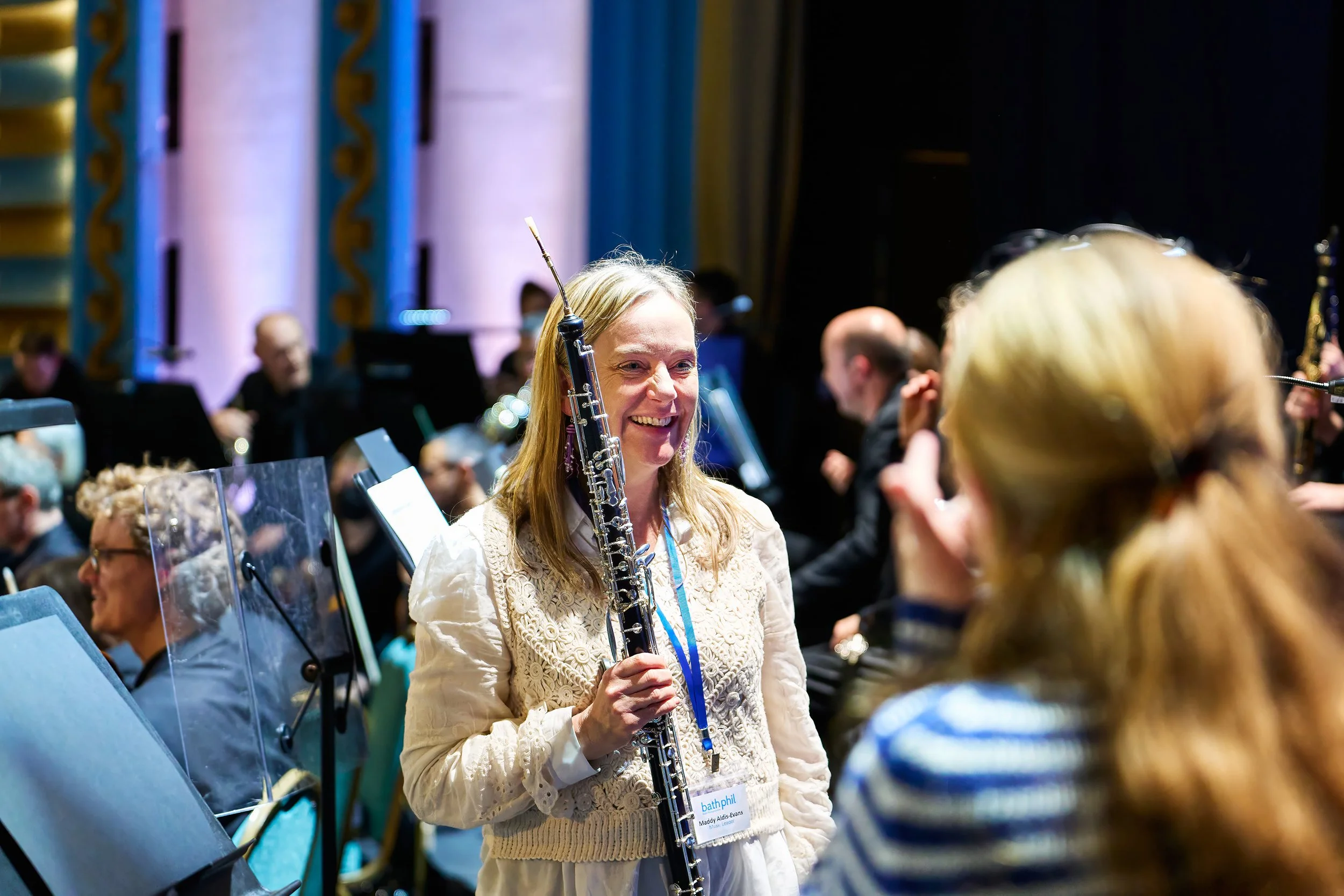 A woman with long blond hair smiling while holding a clarinet at an indoor concert or event, surrounded by other musicians, some playing instruments, in a decorated hall with blue and gold accents.
