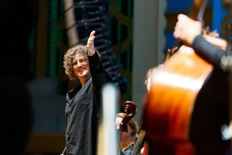 A woman with curly hair waving while standing among musicians playing instruments, possibly in a concert or rehearsal setting.
