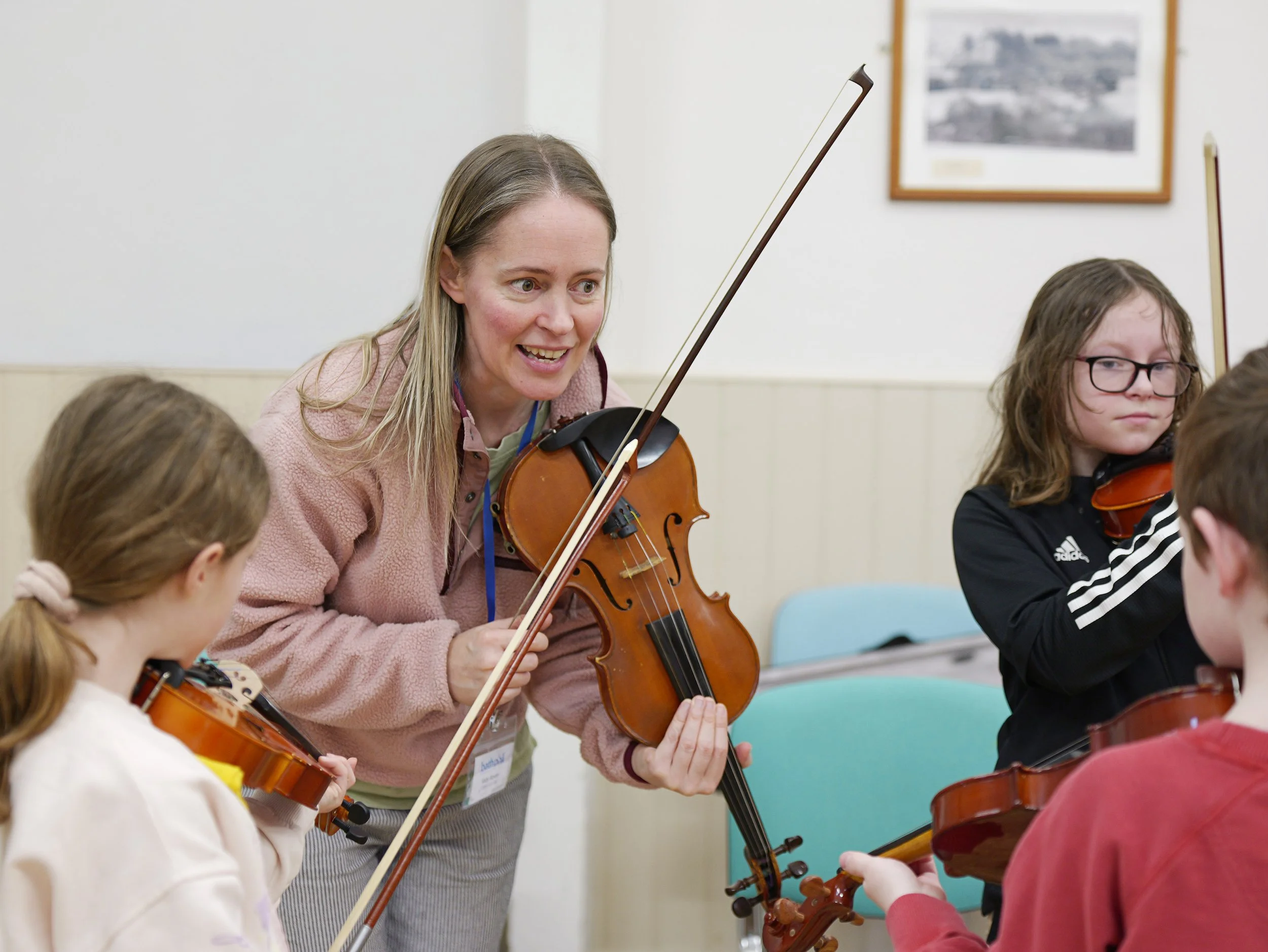 A woman with long blonde hair playing a violin while children with violins listen during a music lesson in a classroom.