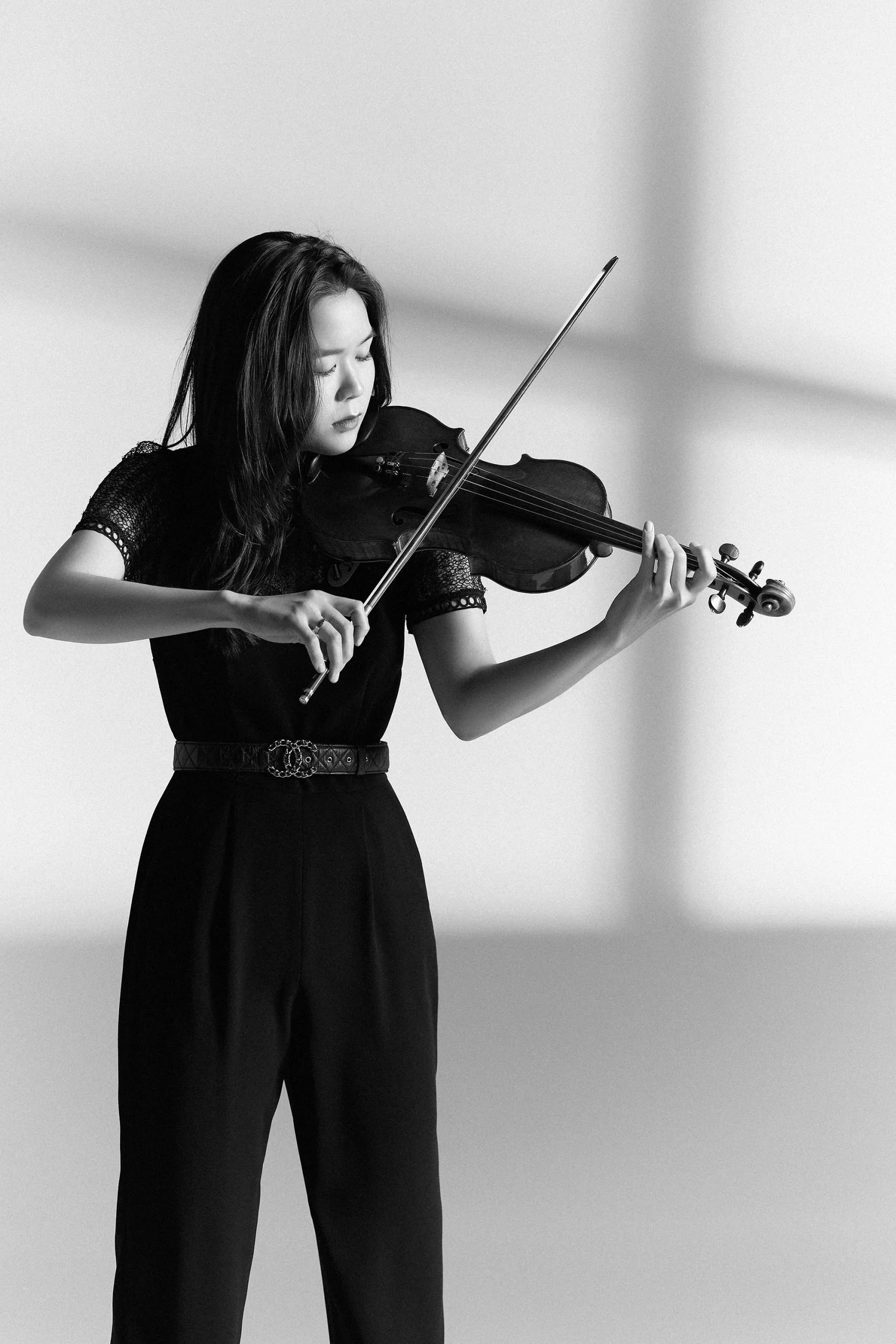 A woman with long dark hair playing a violin in a plain room, dressed in black with a patterned belt, captured in black and white.