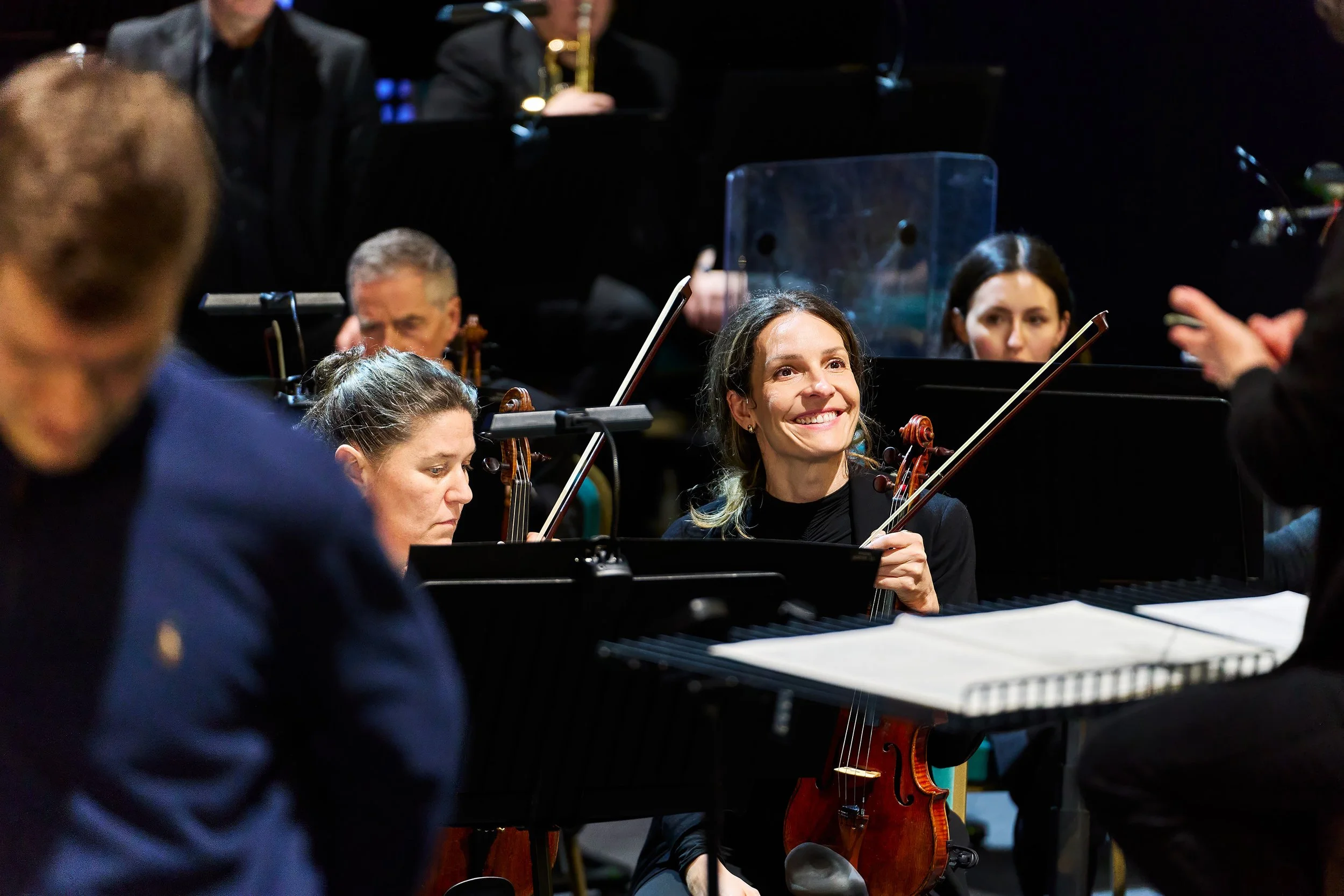 A group of musicians, including women and a man, playing string instruments and a piano in a concert setting, with a woman smiling.