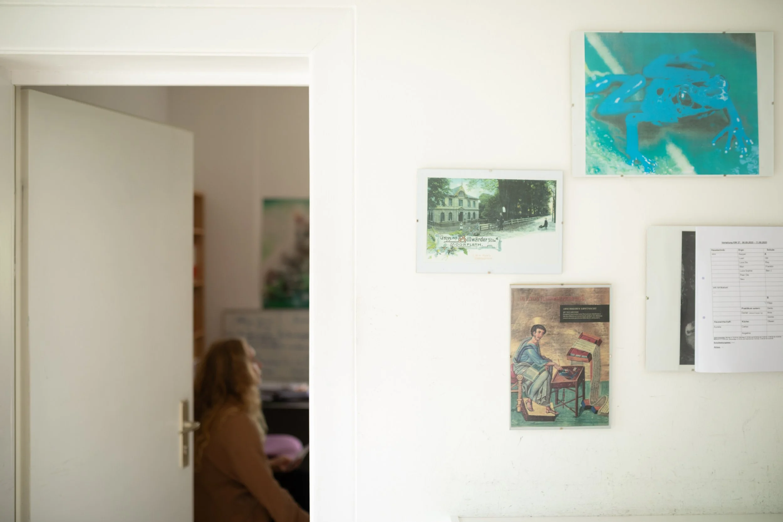 Ein Blick in eine Kunstgalerie mit verschiedenen Gemälden an der Wand. Im Vordergrund eines Mädchens mit roten Haaren, das auf einem Stuhl sitzt und ein Buch liest.