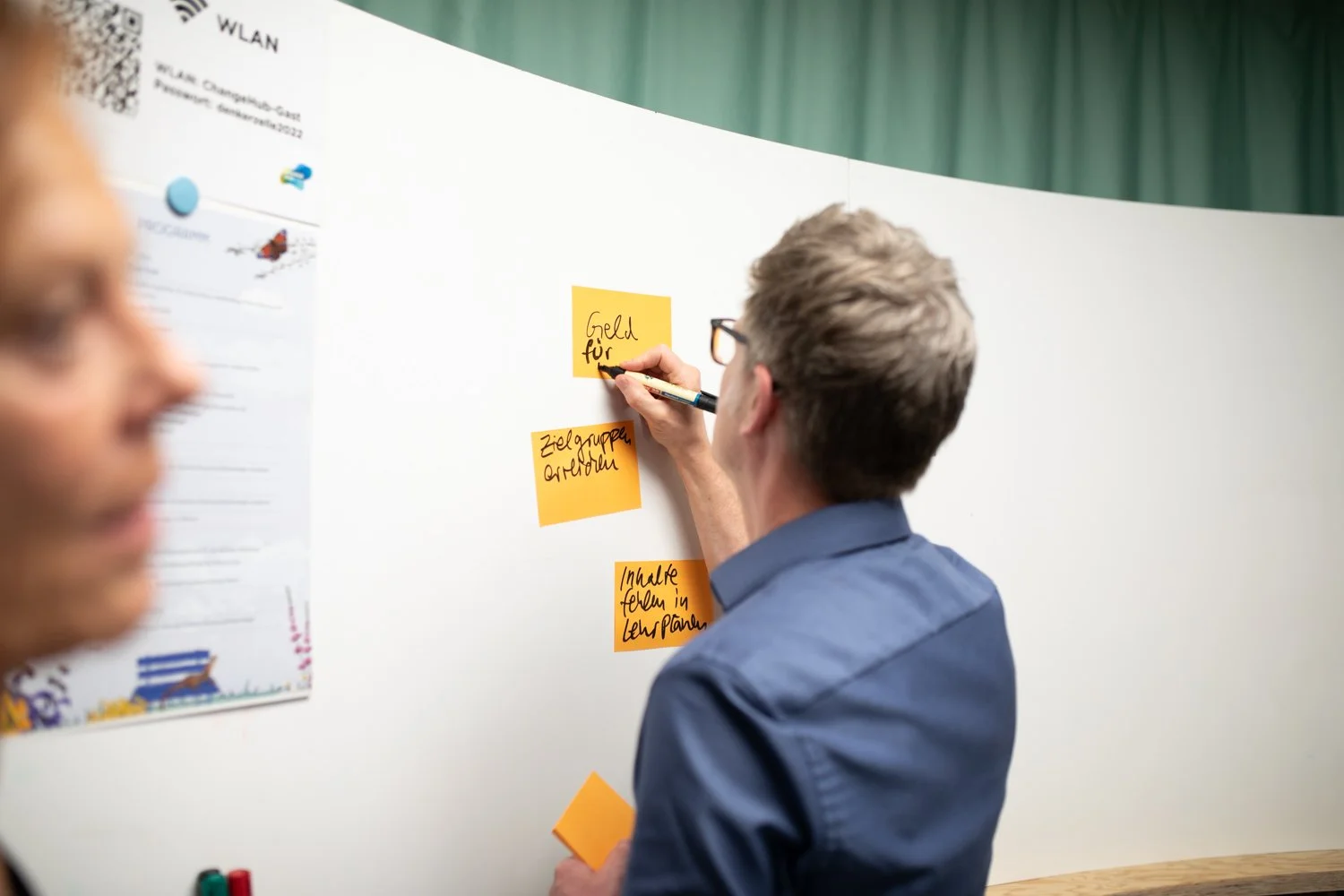 Mann schreibt auf Sticky Notes an einer weißen Wand, im Hintergrund grüne Vorhang. Neben ihm liegen weitere Sticky Notes auf einem Tisch. Es ist eine Besprechung oder ein Brainstorming.