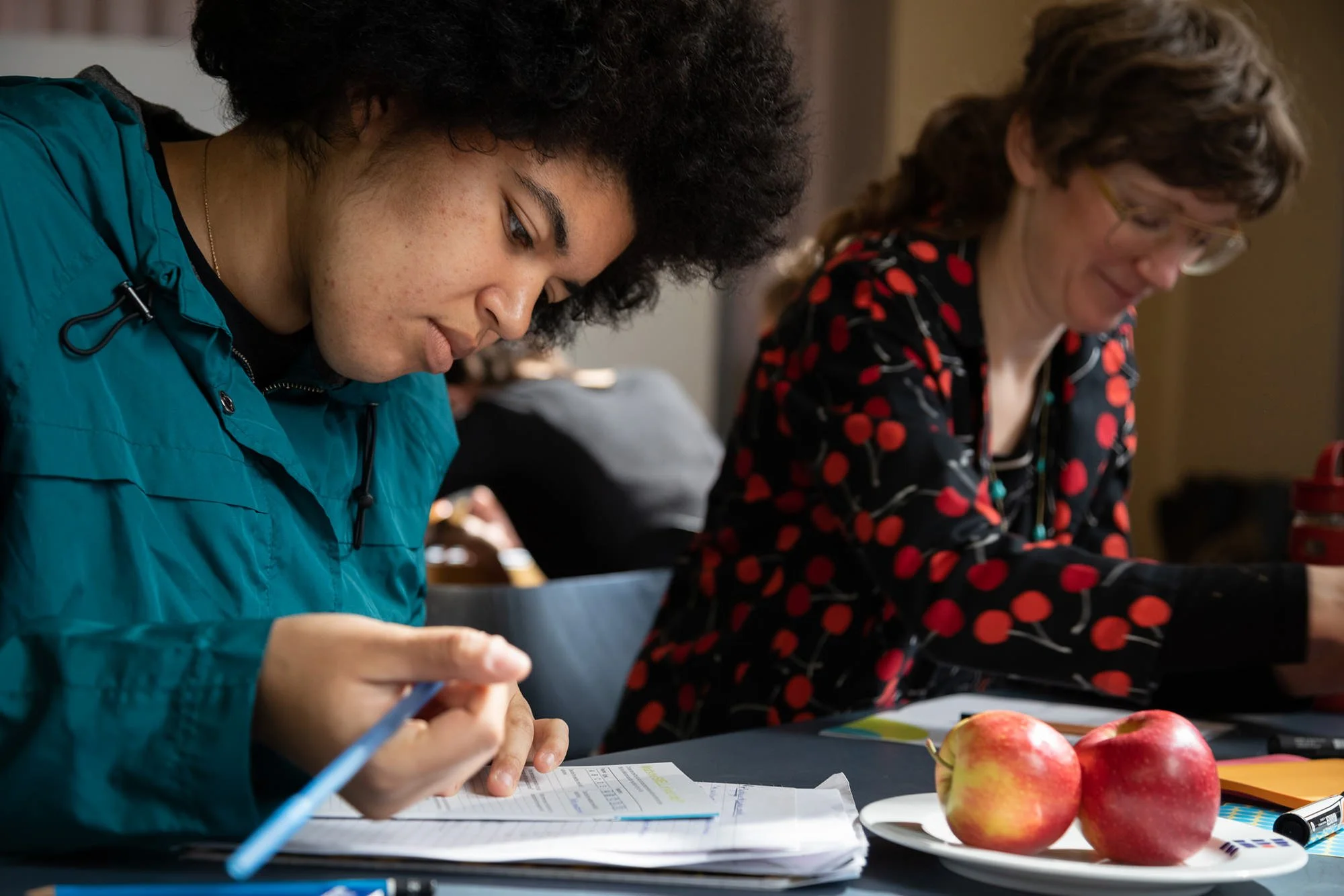 Zwei Frauen sitzen bei einem Workshop an einem Tisch, die eine schreibt in Unterlagen, während die andere nach unten schaut. Auf dem Tisch liegen Äpfel, Dokumente und Schreibutensilien.
