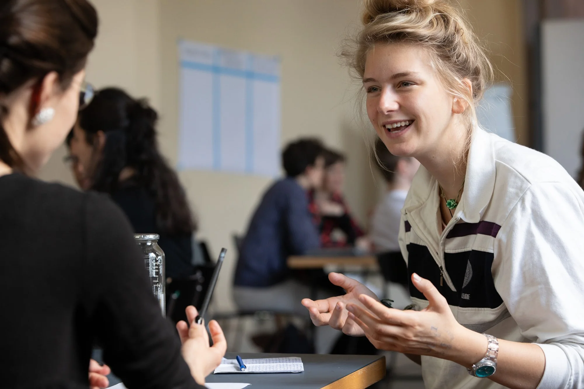 Zwei Frauen, vermutlich Studentinnen, unterhalten sich bei einem Workshop in einem Klassenzimmer. Eine steht lachend und gestikulierend, während die andere zuhört.