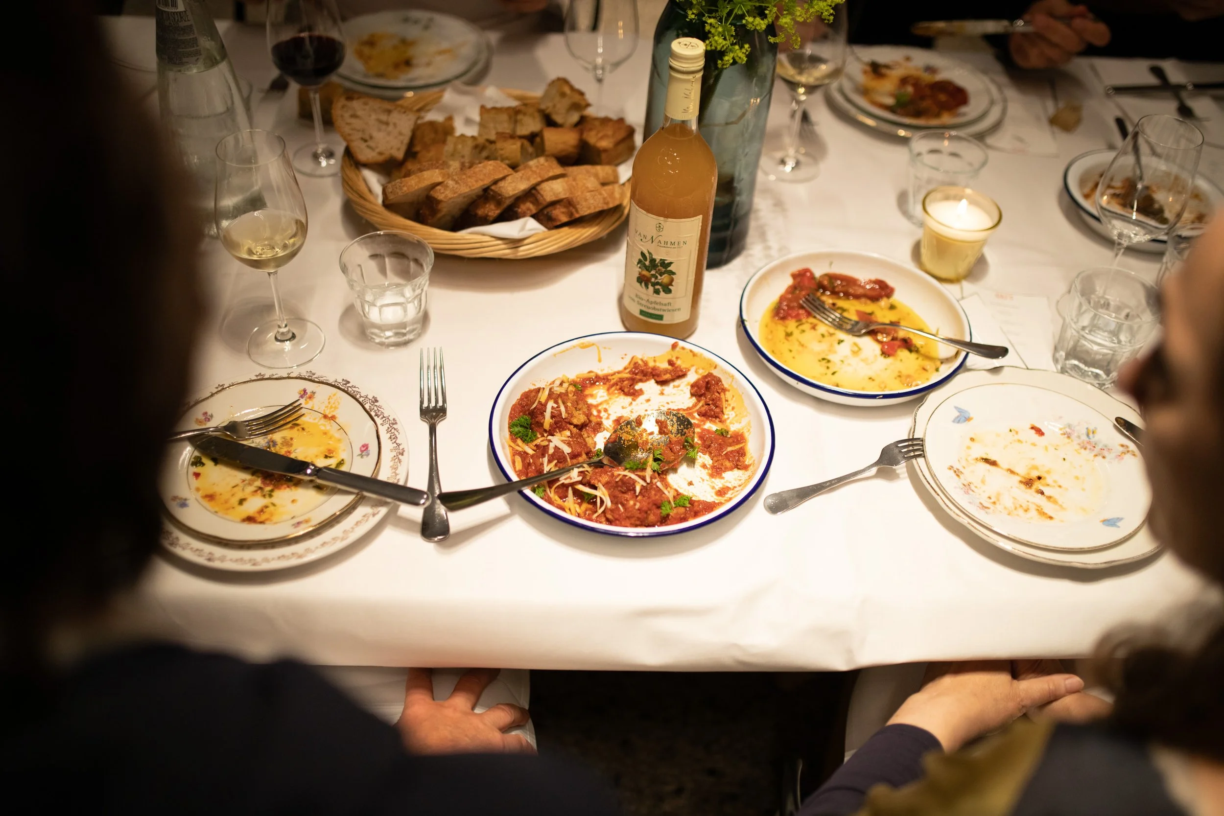 Ein gedeckter Esstisch mit leeren Tellern, Gläsern mit Wein, einem Korb mit Brot, mehreren Schüsseln mit italienischen Gerichten und einer Flasche Wein in einem festlichen Rahmen.