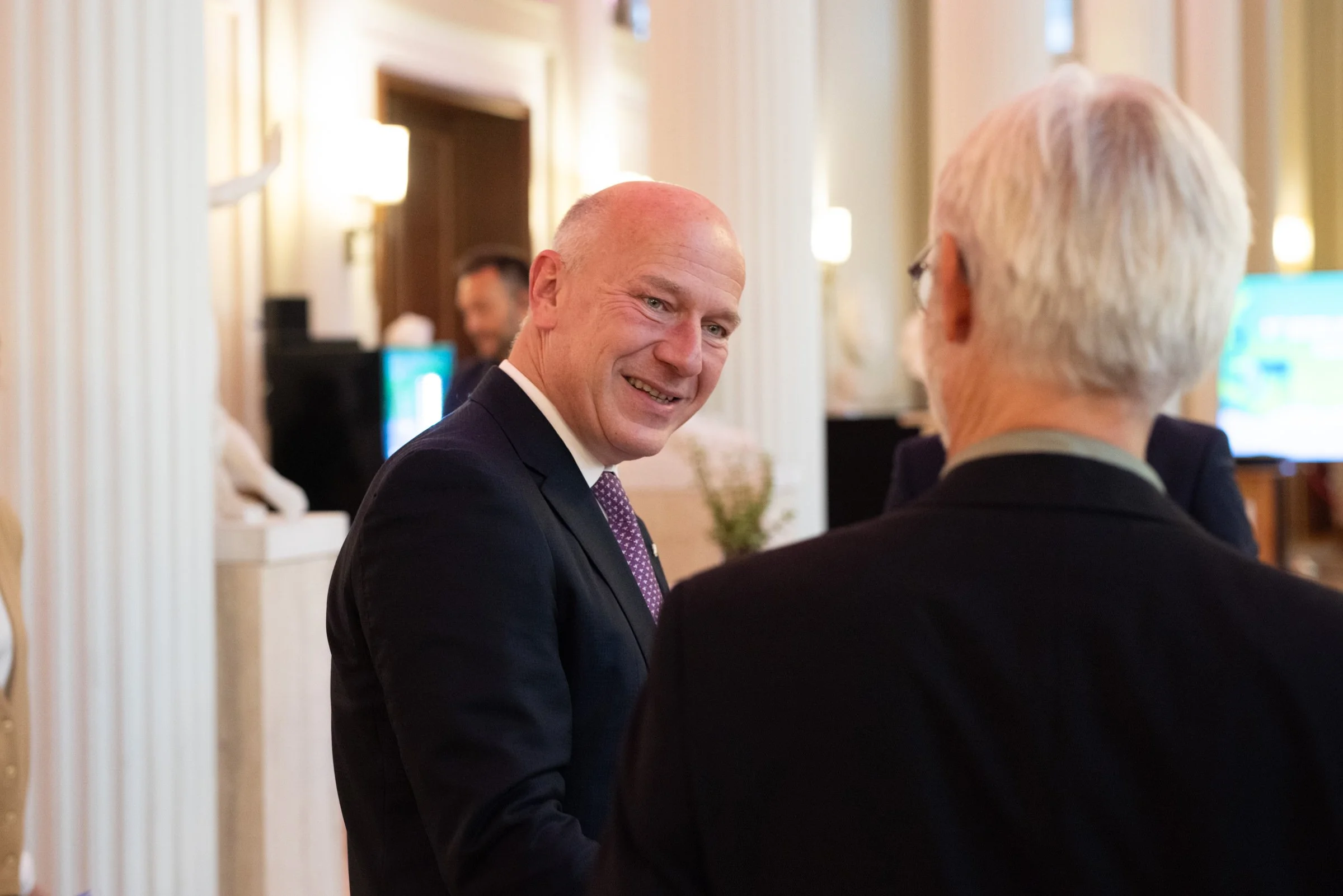 Zwei Männer im Gespräch in einem eleganten Raum mit weicher Beleuchtung. Der eine Mann ist der Regierende Bürgermeister von Berlin, Kai Wegner. Er trägt einen dunklen Anzug und hat eine Glatze, der andere ist von hinten sichtbar und hat weiße Haare.