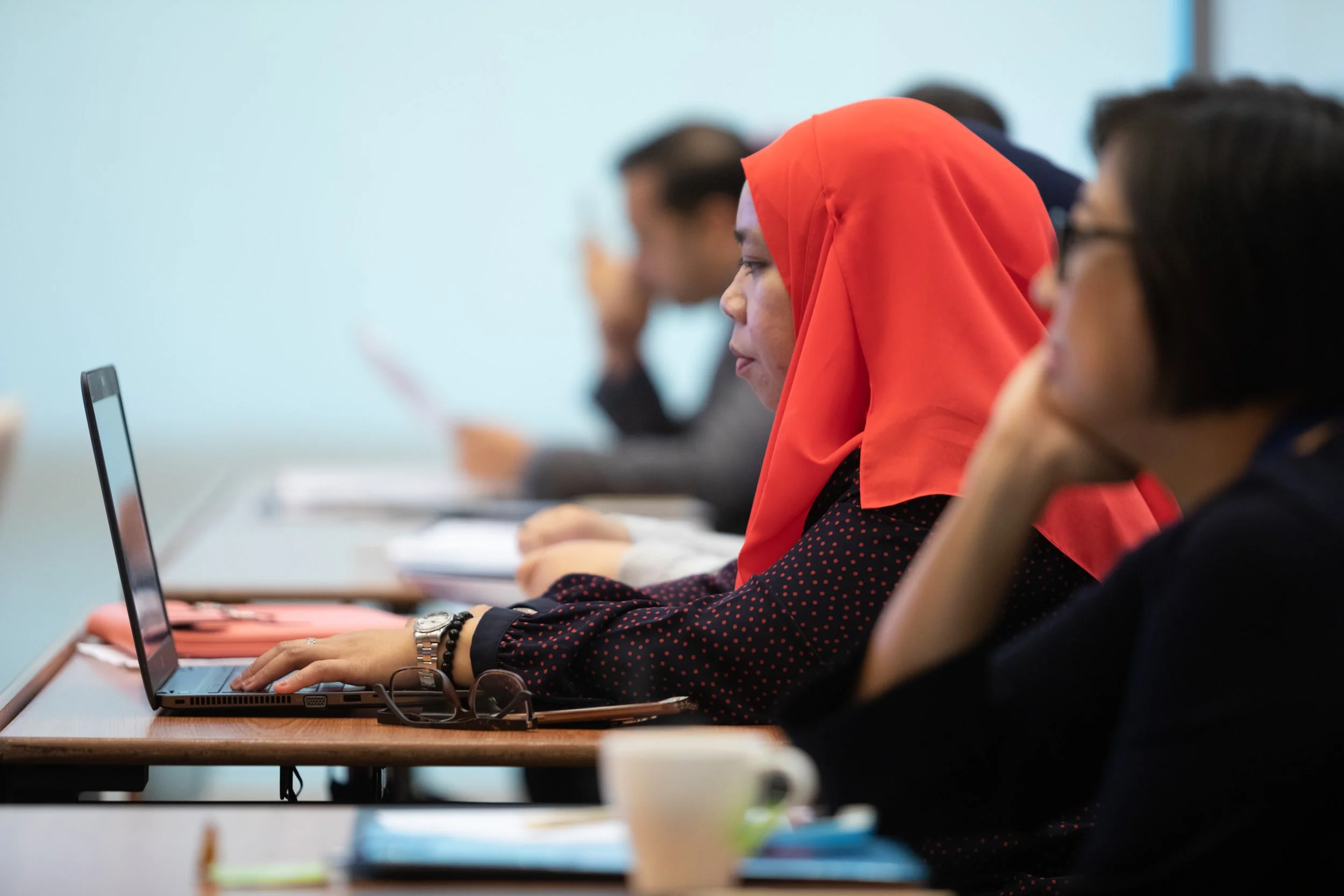 Menschen sitzen an Tischen in einem Klassenraum oder Seminarraum, arbeiten an Laptops. Eine Frau trägt eine rote Kopfbedeckung.