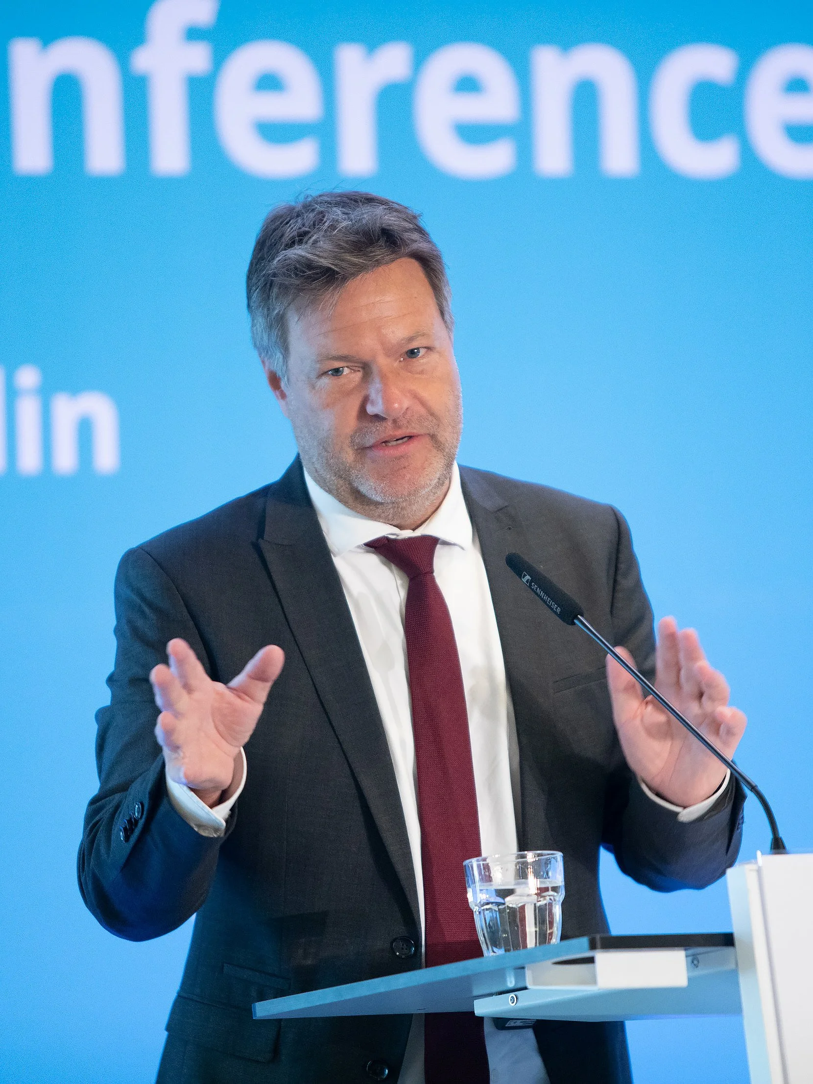 Ein Mann in Anzug und Krawatte spricht bei einer Pressekonferenz, vor einem blauen Hintergrund mit weißen Buchstaben, gestikuliert mit den Händen, steht hinter einem Pult mit einem Glas Wasser und einem Mikrofon.