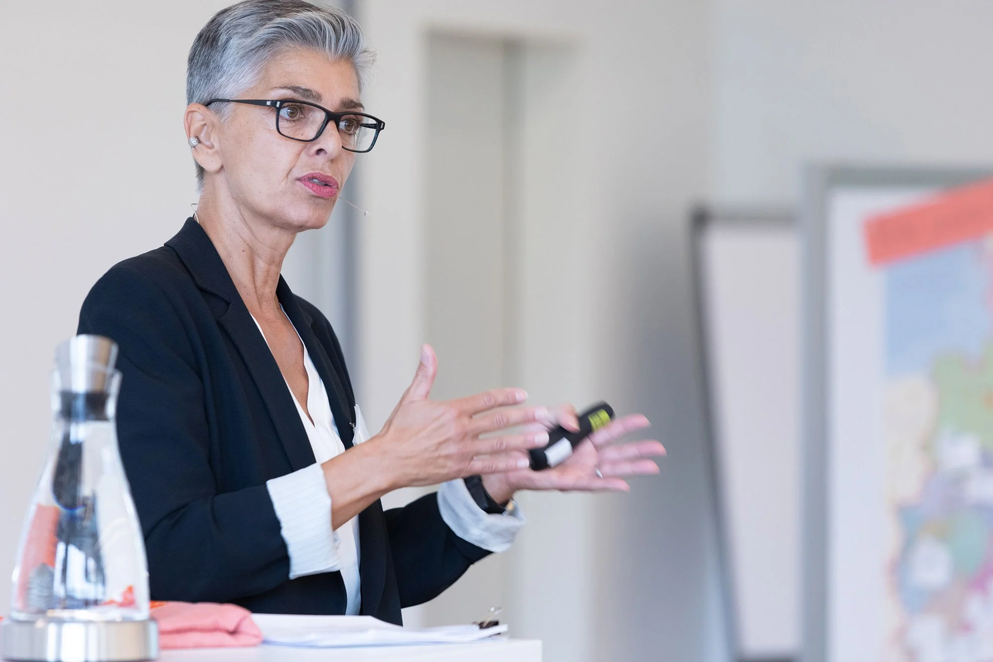 Eine ältere Frau mit kurzen grauen Haaren, schwarzen Brillen und einem schwarzen Blazer präsentiert einen Button oder ein Gerät während einer Präsentation. Sie hat ein Mikrofon am Ohr und sitzt an einem weißen Tisch, auf dem sich eine Wasserflasche u