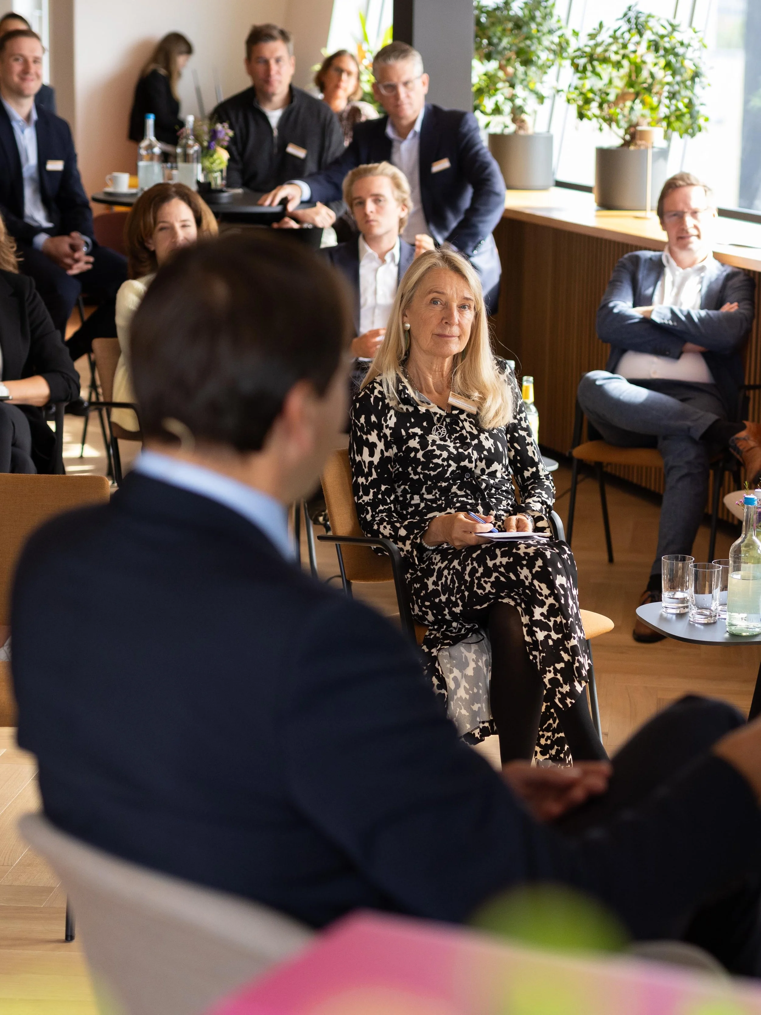 Mehrere Menschen sitzen in einem Raum bei einer Besprechung oder Konferenz. Im Vordergrund ist eine Frau mit blondem Haar in einem schwarzen und weißen Kleid zu sehen, die Notizen macht. Im Hintergrund sitzen weitere Personen, die aufmerksam sind, un