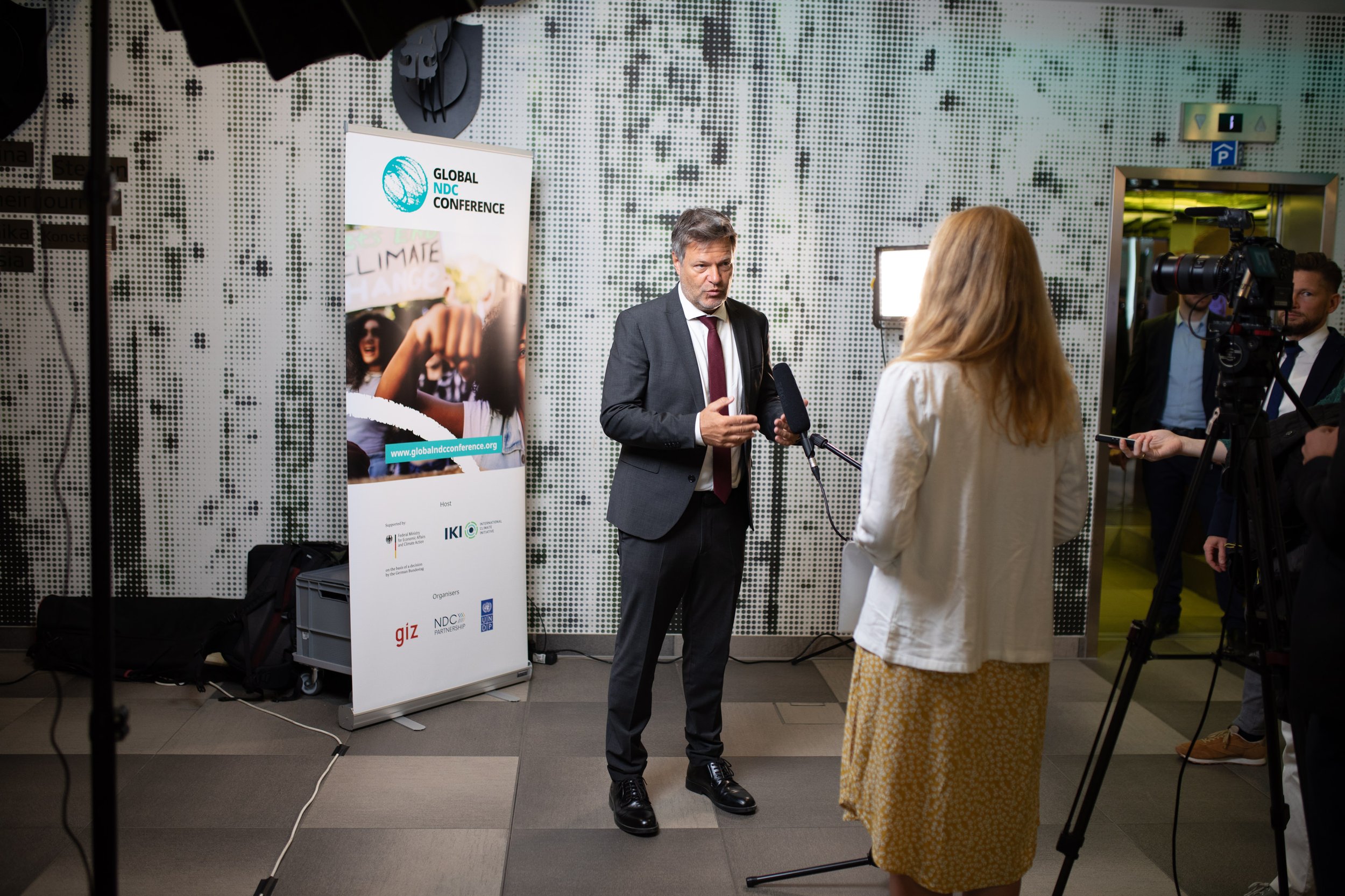 Mann in Anzug im Interview mit Frau in gelbem Rock bei der Konferenz. Kamera filmt, Banner und Plakate im Hintergrund.