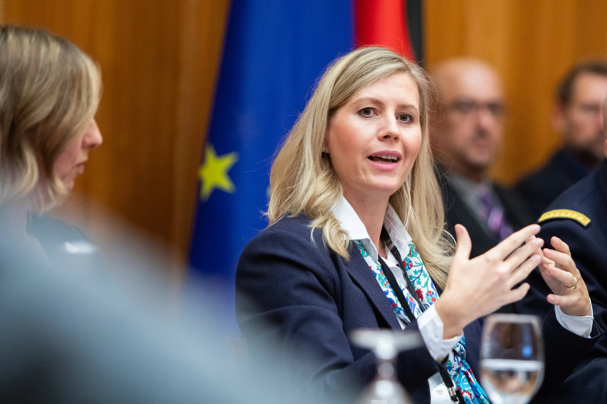 Eine blonde Frau spricht bei einer offiziellen Veranstaltung, im Hintergrund Flaggen der Europäischen Union und eines anderen Landes.