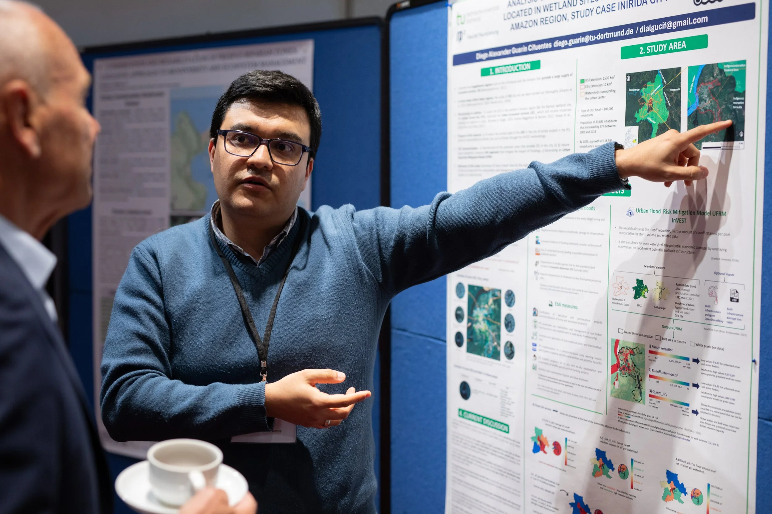 Zwei Männer diskutieren vor einem wissenschaftlichen Poster auf einer Konferenz. Einer erklärt das Poster, das Karte und Diagramme enthält, während der andere zuhört, während er eine Tasse hält.