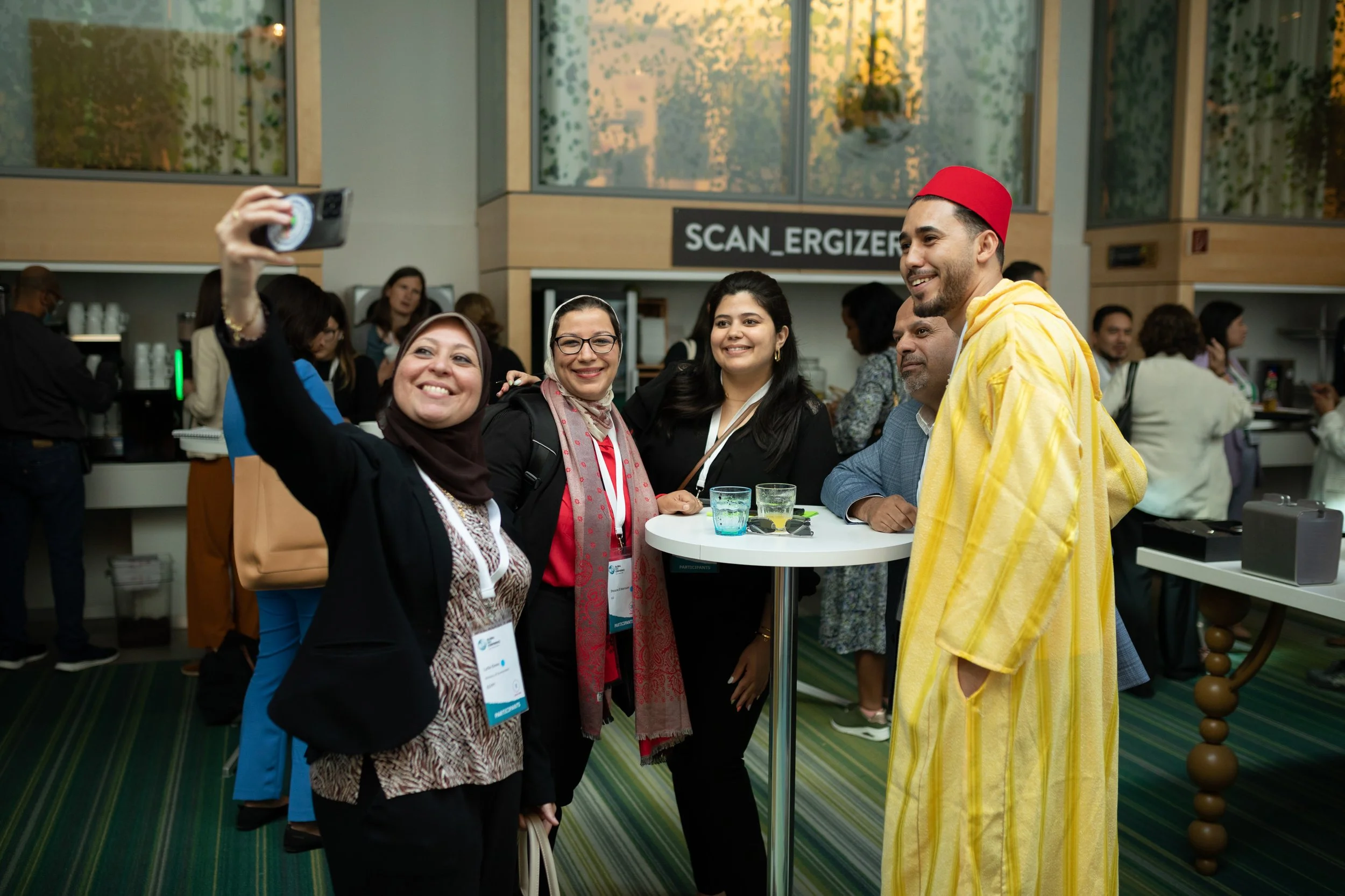 Eine Gruppe von vier Menschen, darunter zwei Frauen und zwei Männer, posieren für ein Selfie in einer modernen Veranstaltungsumgebung. Eine Frau trägt einen Hijab, eine andere trägt eine Brille und einen Schal. Der Mann trägt eine rote traditionelle 