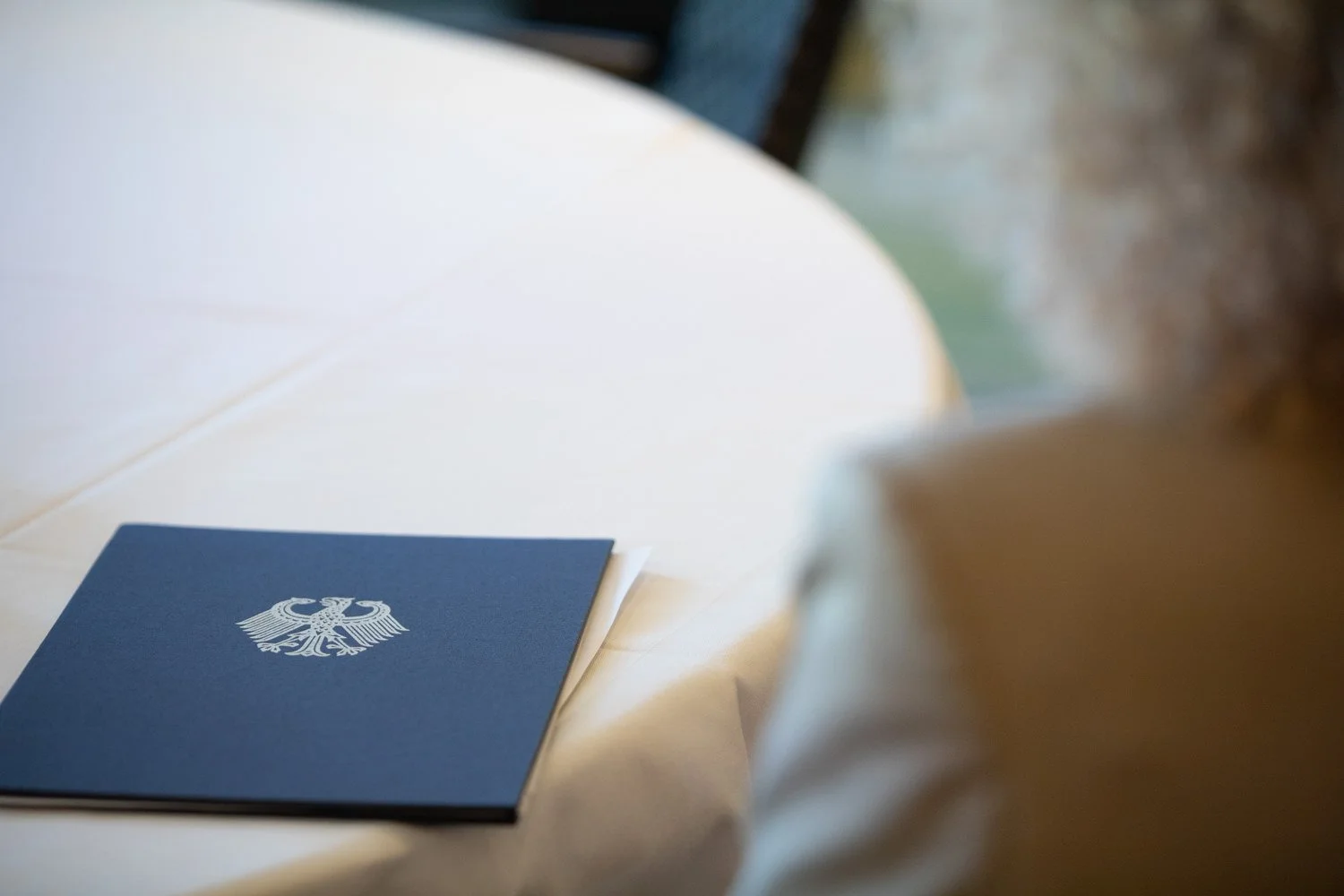 Ein blauer Ordner mit dem Bundesadler-Emblem liegt auf einem weißen Tischtuch.