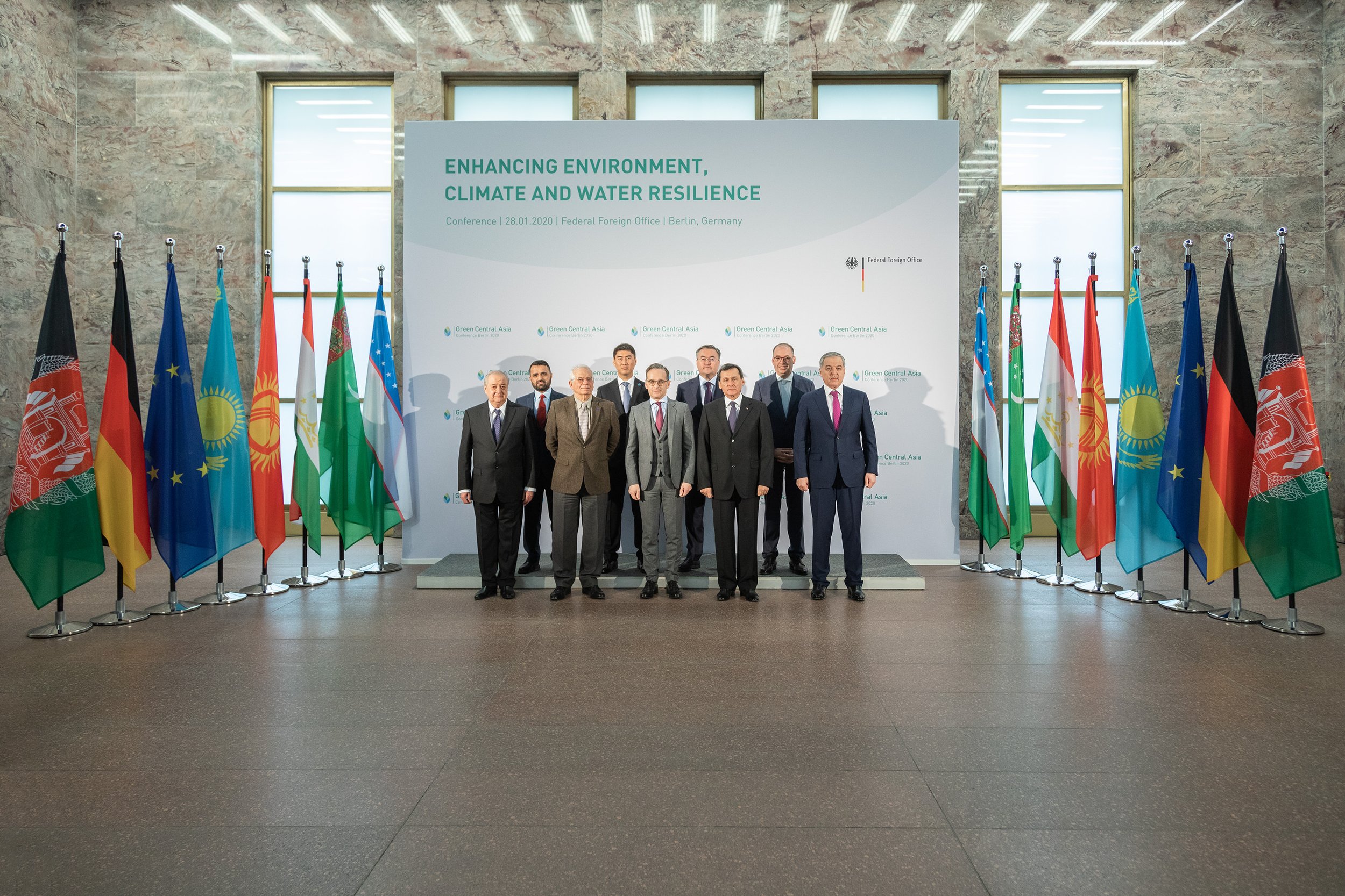 Gruppenfoto von mehreren Männern in formeller Kleidung vor einer großen Plakatwand bei einer Konferenz in Berlin, Deutschland, mit einer Reihe von internationalen Flaggen im Hintergrund.