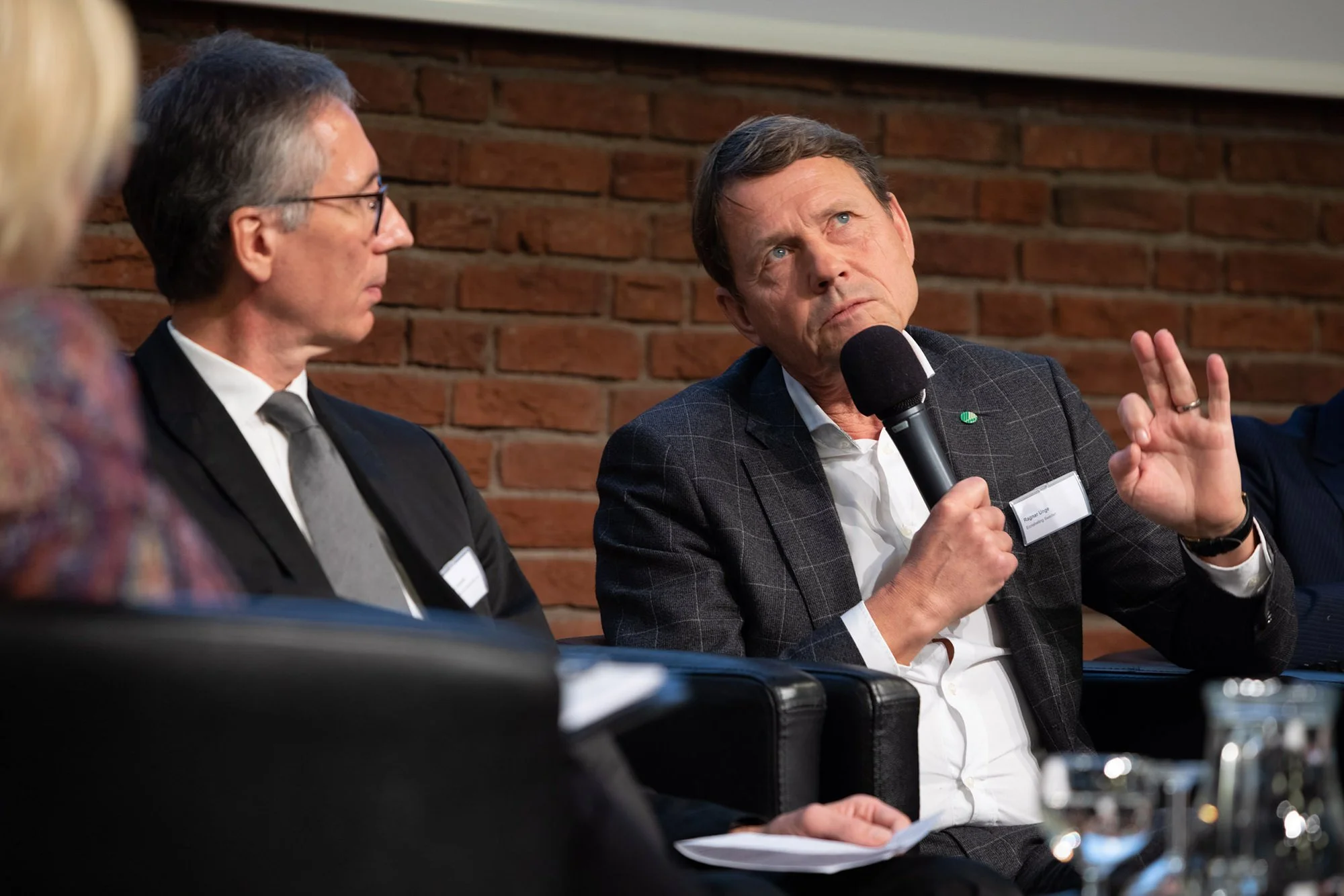 Ein Mann im Anzug spricht mit einem Mikrofon bei einer Diskussion, während zwei weitere Personen zuhören. Die Szene findet in einem Raum mit einer roten Backsteinwand statt.