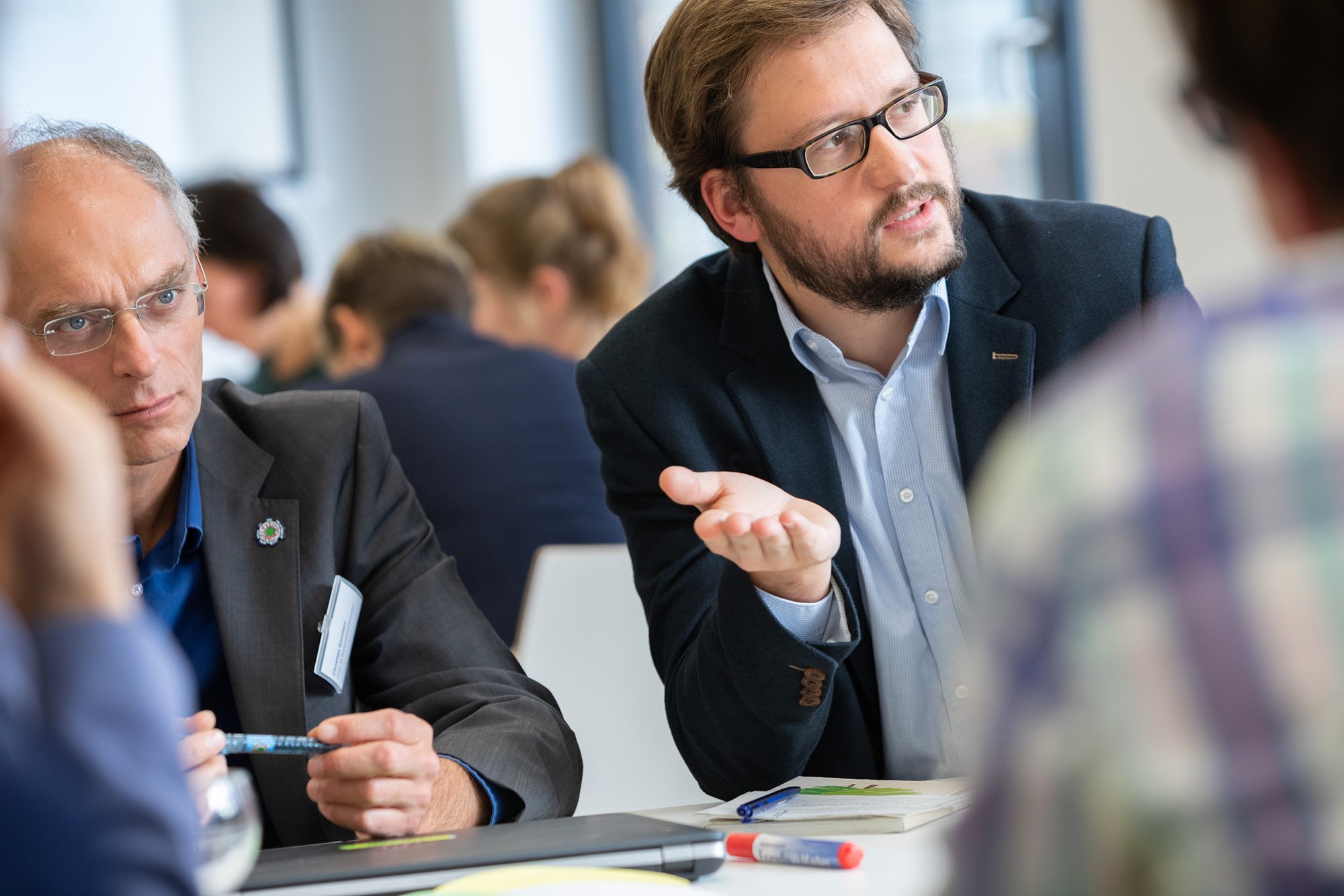 Ein Mann mit Brille und Bart sitzt an einem Tisch, spricht und gestikuliert mit den Händen. Im Hintergrund sind weitere Menschen in einem Sitzungsraum an einem Meeting beteiligt.