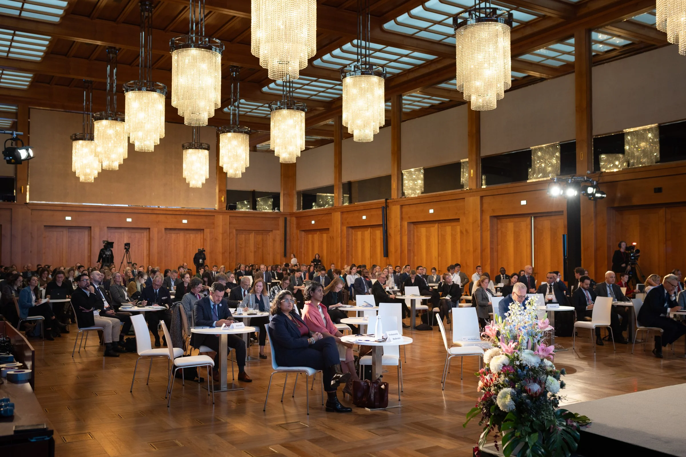 Innenansicht eines großen Raums während einer Konferenz mit vielen anwesenden Menschen, prunkvollen Kronleuchtern an der Decke und einem Blumenarrangement auf der Bühne.