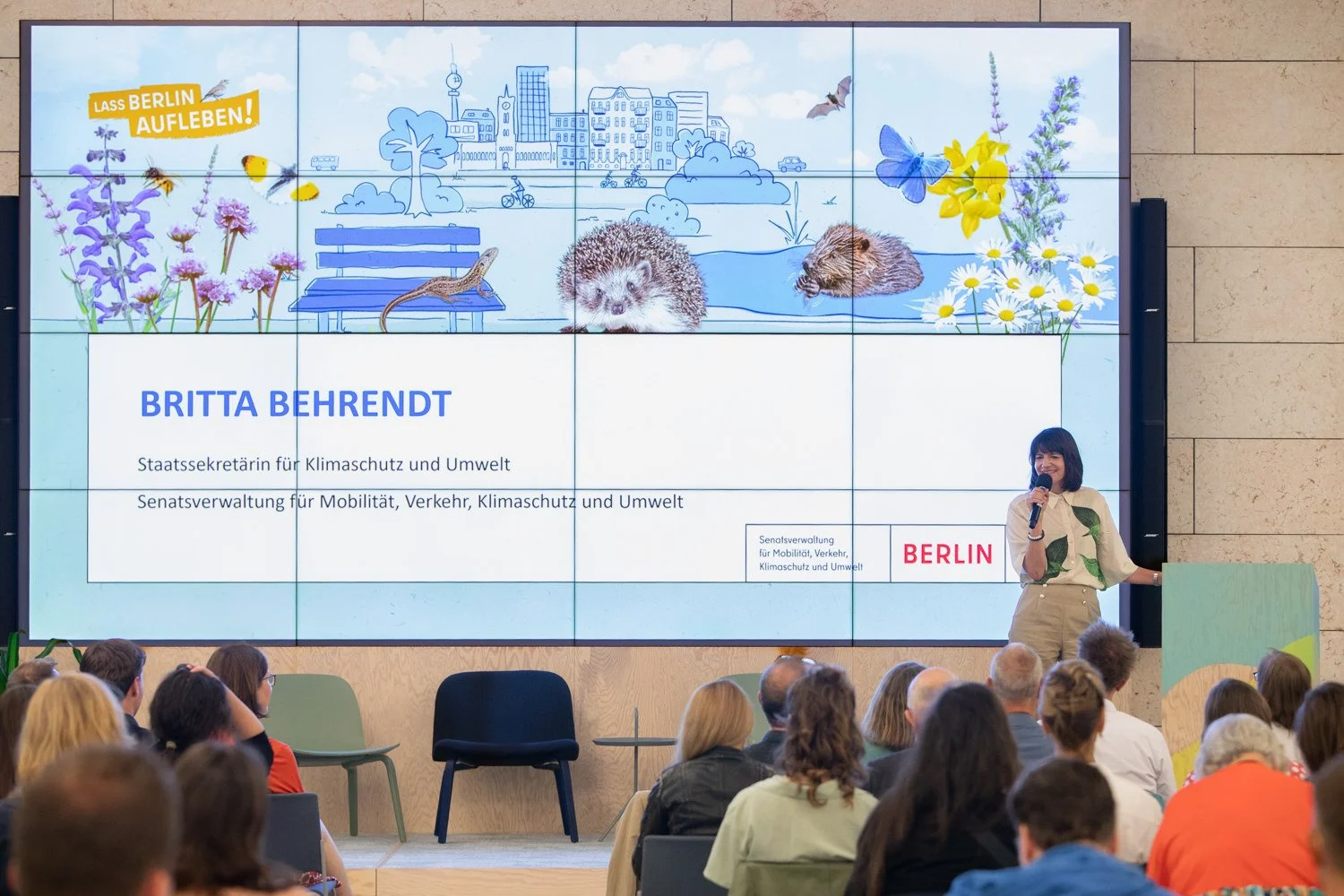 Frau hält eine Rede vor Publikum in einem Veranstaltungsraum mit großem Bildschirm im Hintergrund, der eine Grafik von Berliner Stadtansichten und Natur zeigt. Text auf dem Bildschirm: "Britta Behrendt, Staatssekretärin für Klimaschutz und Umwelt, Se