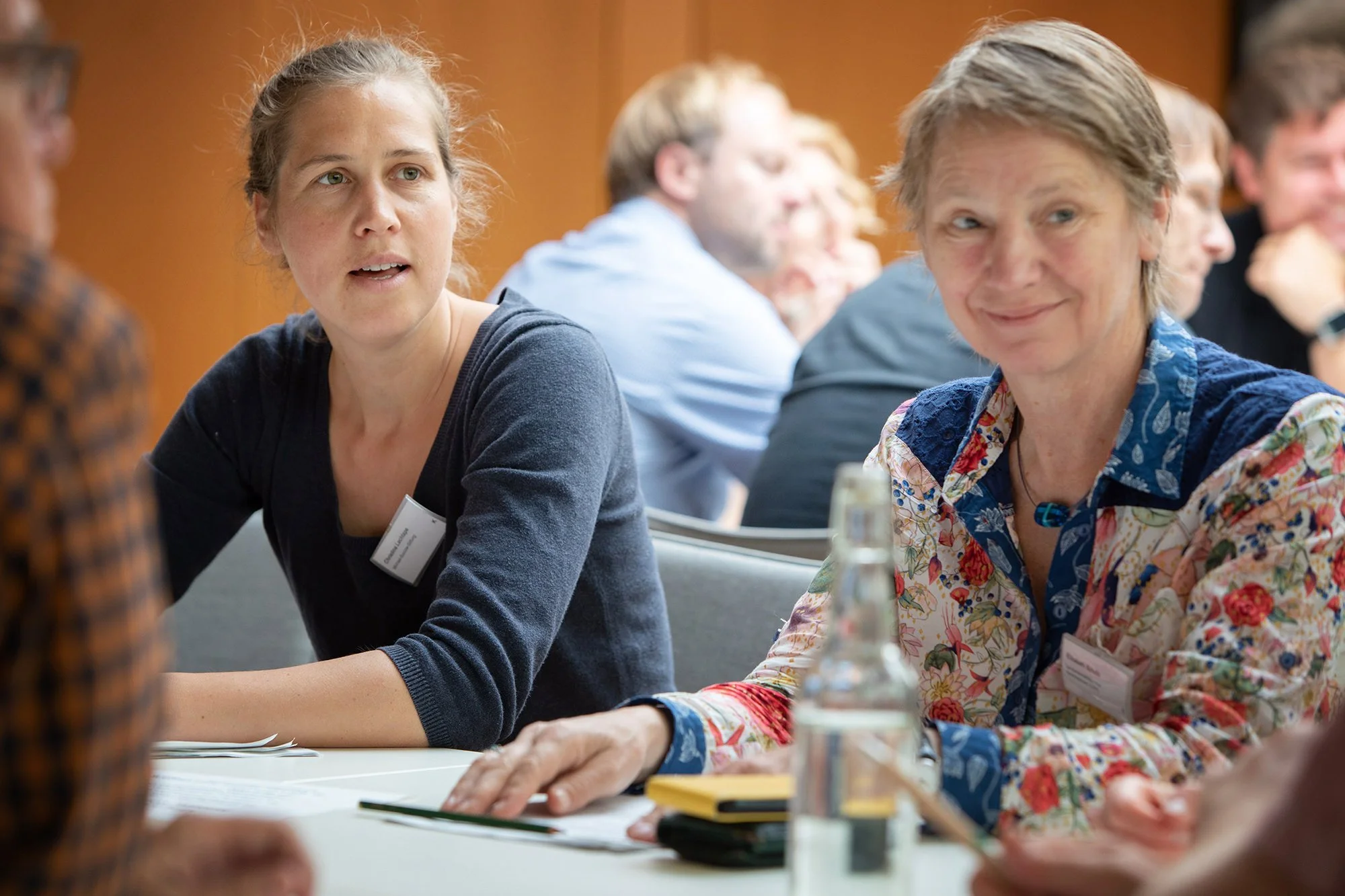 Zwei Frauen sitzen an einem Tisch im Gespräch in einem Raum mit weiteren Personen im Hintergrund, im Vordergrund ein Glas Mineralwasser und Schreibmaterial sichtbar.