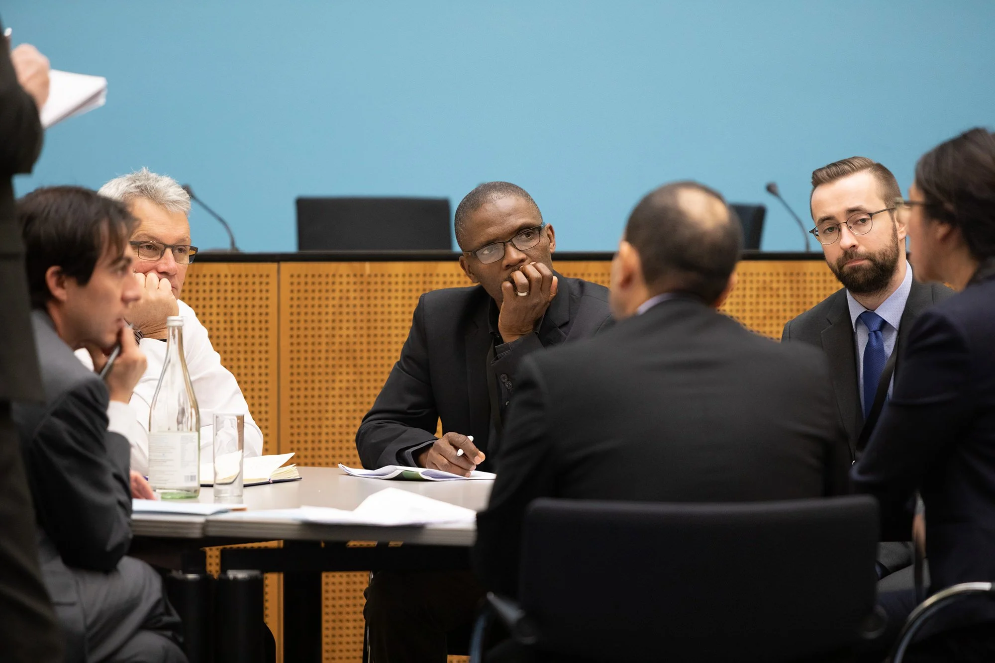 Fünf Menschen sitzen an einem runden Tisch bei einer Besprechung oder Diskussion, in einem Raum mit blauer Wand und Mikrofonen auf dem Tisch.