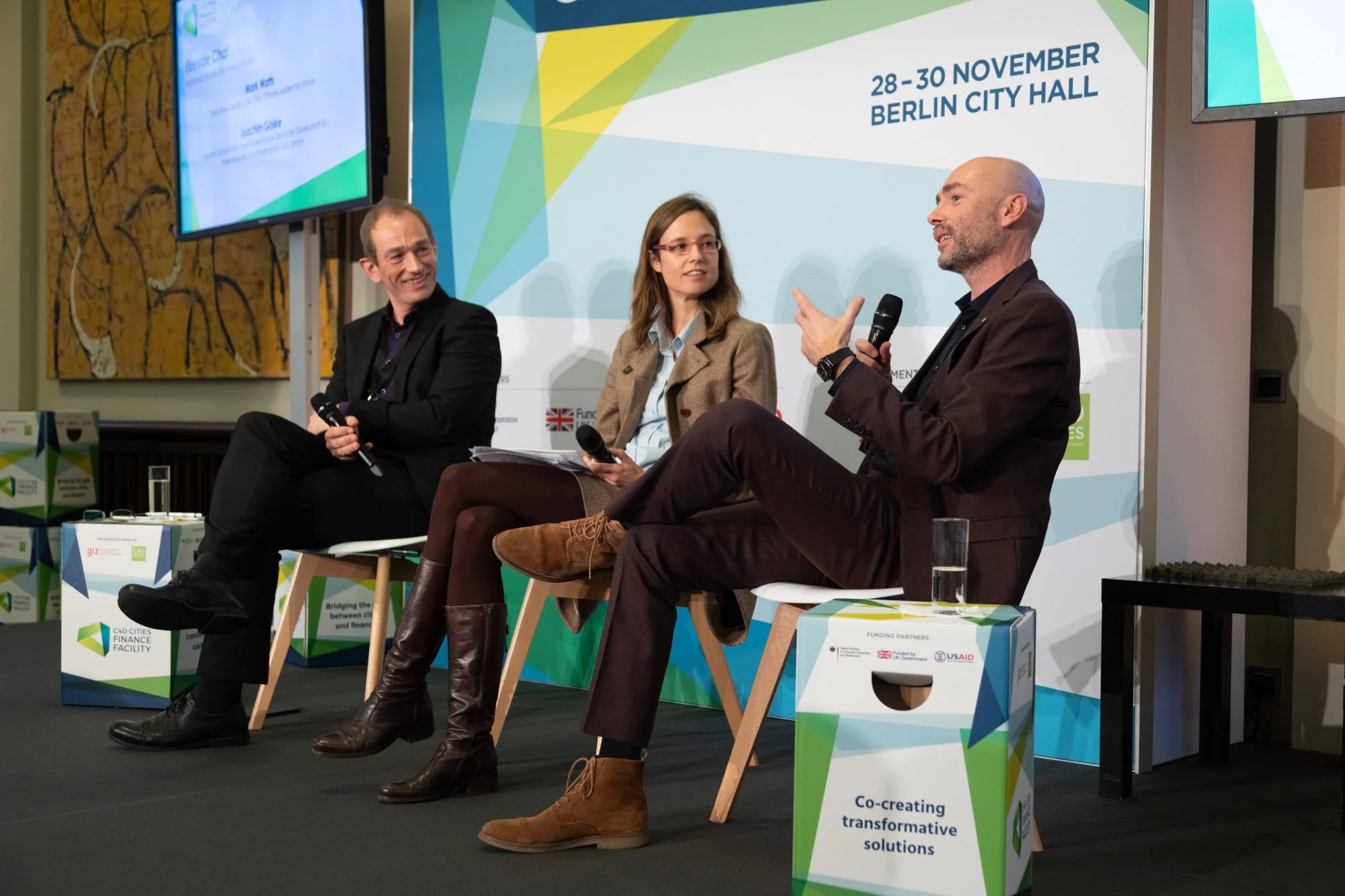 Drei Personen sitzen auf Stühlen bei einer Diskussion während eines Konferenz-Events im Berliner Rathaus. Im Hintergrund ein digitaler Bildschirm mit Veranstaltungstext und eine große Veranstaltungsankündigung mit Daten, Ort und Partnern, darunter UK
