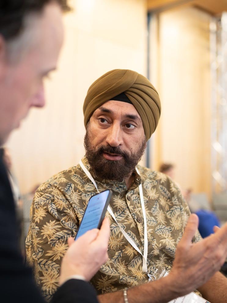 Ein Mann mit türkischem Turban und Batik-Hemd spricht mit einer anderen Person, die ein Smartphone hält, in einem hell erleuchteten Raum.