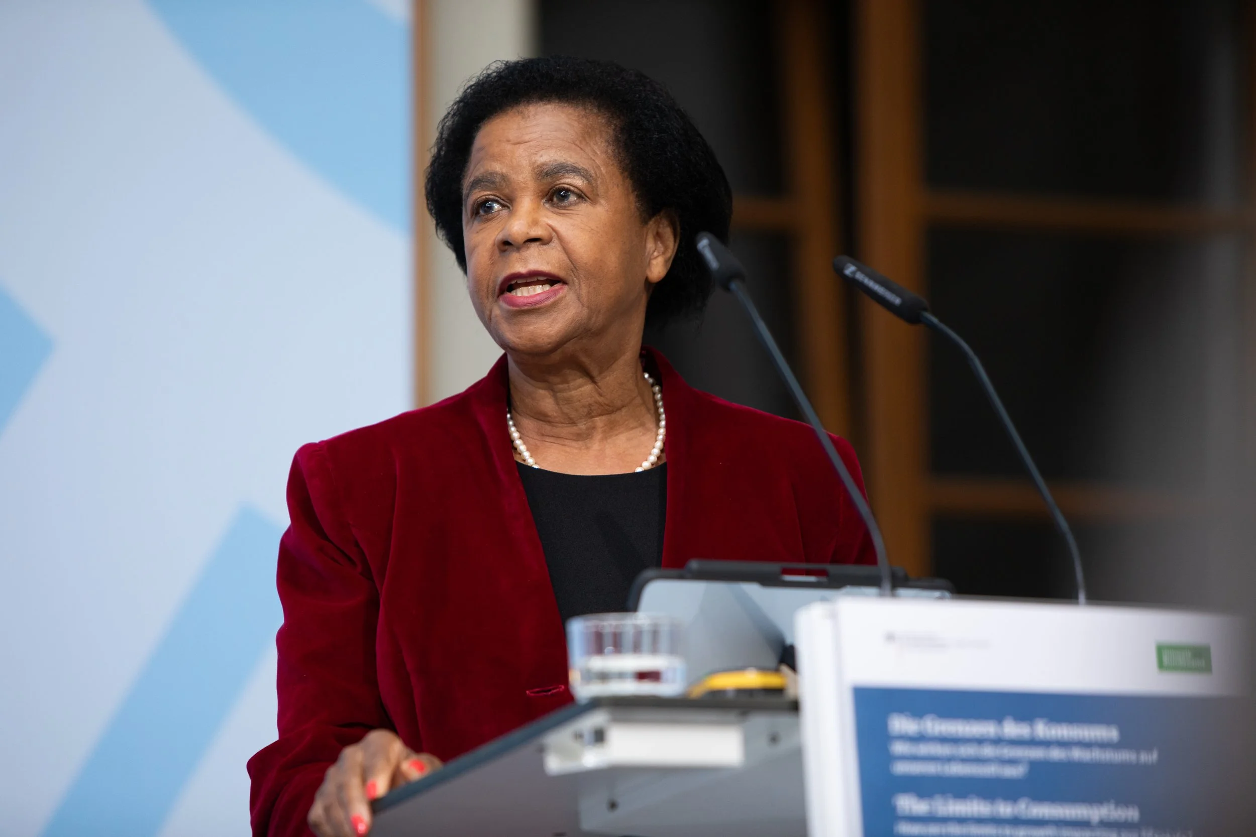 Standbild einer Frau, die an einem Rednerpult spricht, trägt eine rote Jacke und einer Perlenkette, im Hintergrund ein Bildschirm mit Text auf Deutsch.