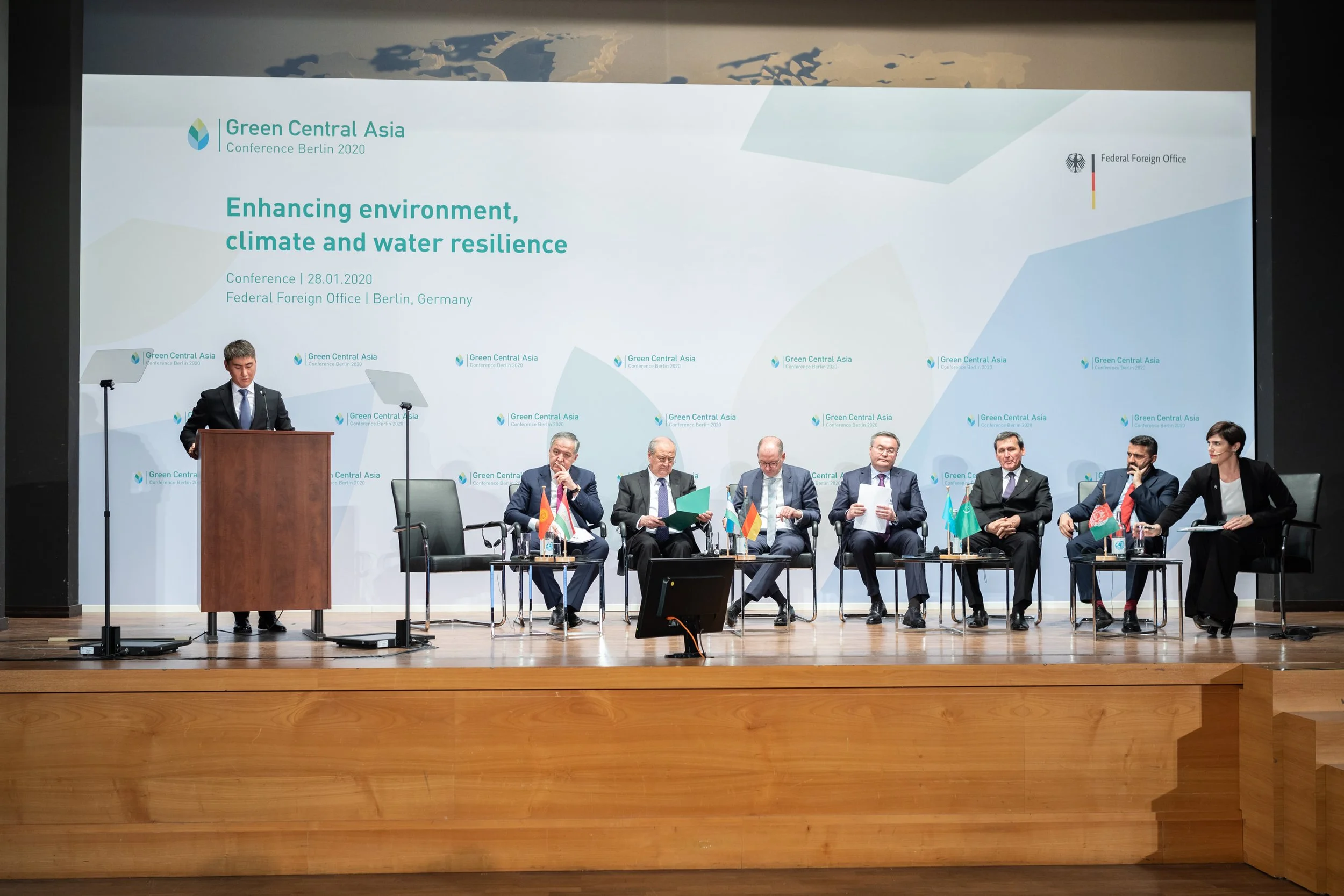 Ein Konferenzsaal mit einer Präsentation auf einer großen Leinwand. Auf der Bühne steht eine Frau am Rednerpult, im Hintergrund sitzen sechs Männer und eine Frau an einem Tisch, alle mit Ländernflaggen vor sich. Das Thema ist Umwelt, Klima und Wasser