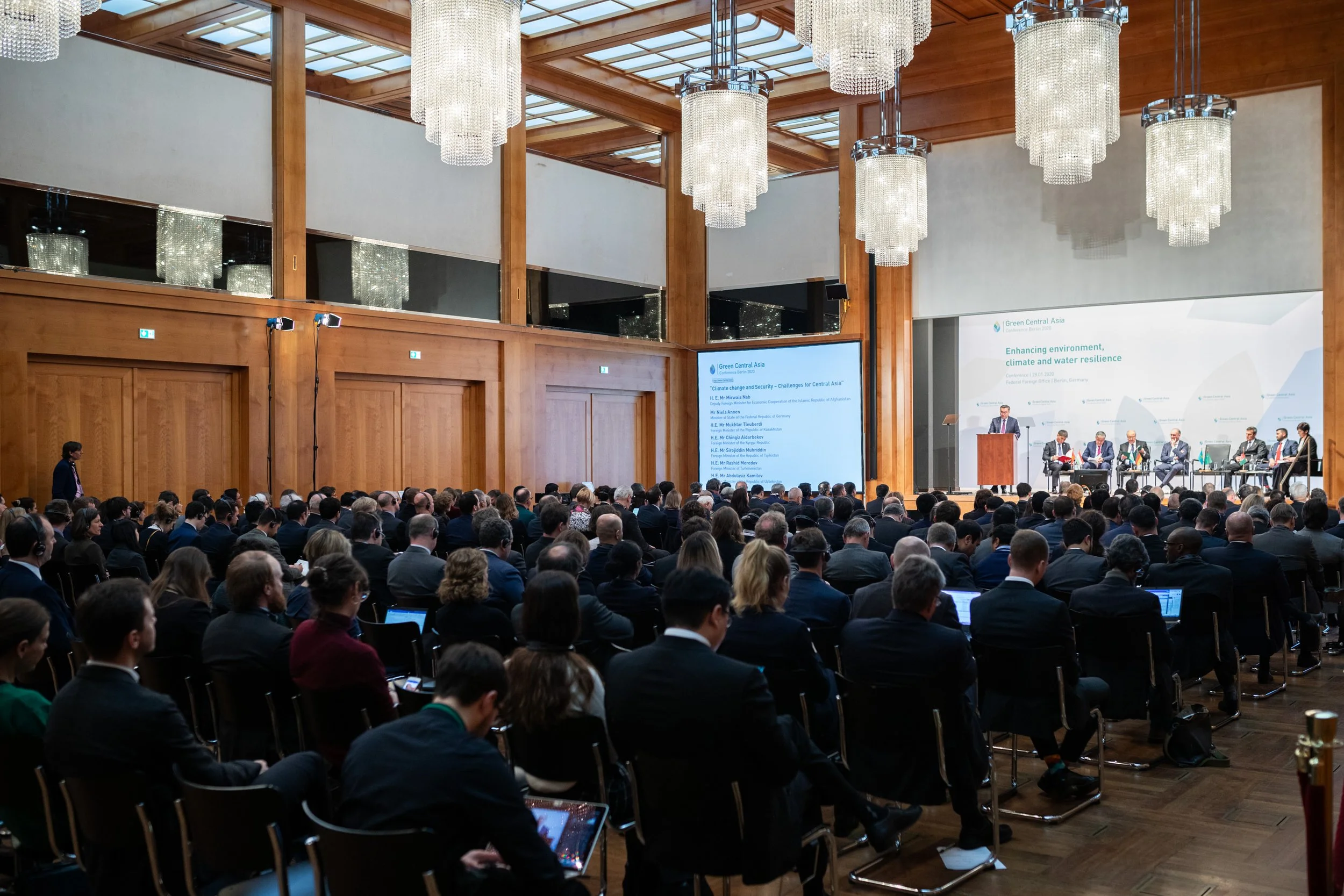 Binnenaufnahme einer großen Konferenzhalle mit vielen Teilnehmern, an einem Podium spricht ein Redner zu einem Publikum, im Hintergrund sind Projektionsbildschirme mit Text zu Umwelt- und Klimafragen.