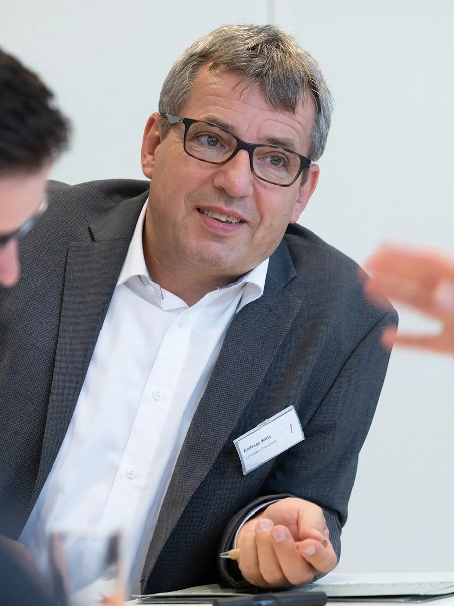 Ein Mann mit Brille in einem Anzug spricht bei einem Treffen, hält einen Stift in der Hand, trägt eine Namensschild, das auf 'Andreas Witte' hinweist.
