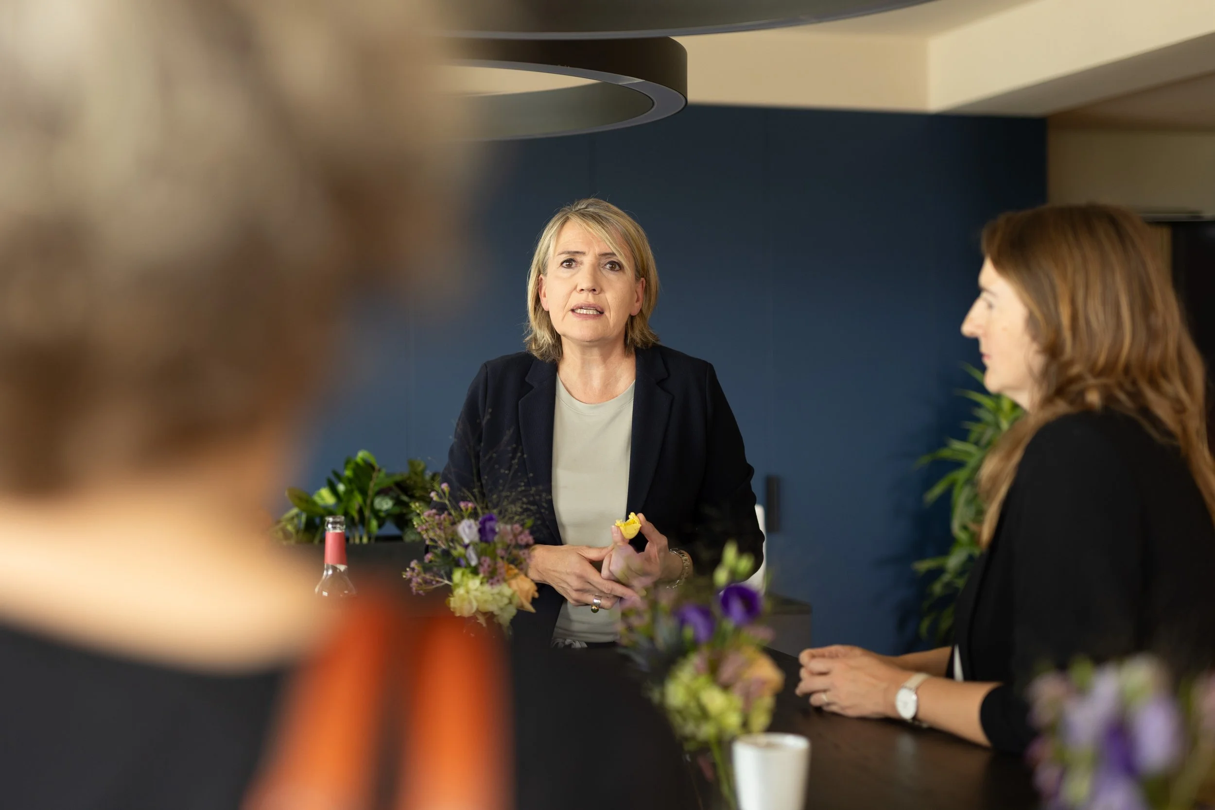 Drei Frauen sitzen an einem Tisch in einem Besprechungsraum, die mittlere Frau spricht, während die anderen beiden zuhören. Der Tisch ist mit Blumen und Getränken dekoriert.