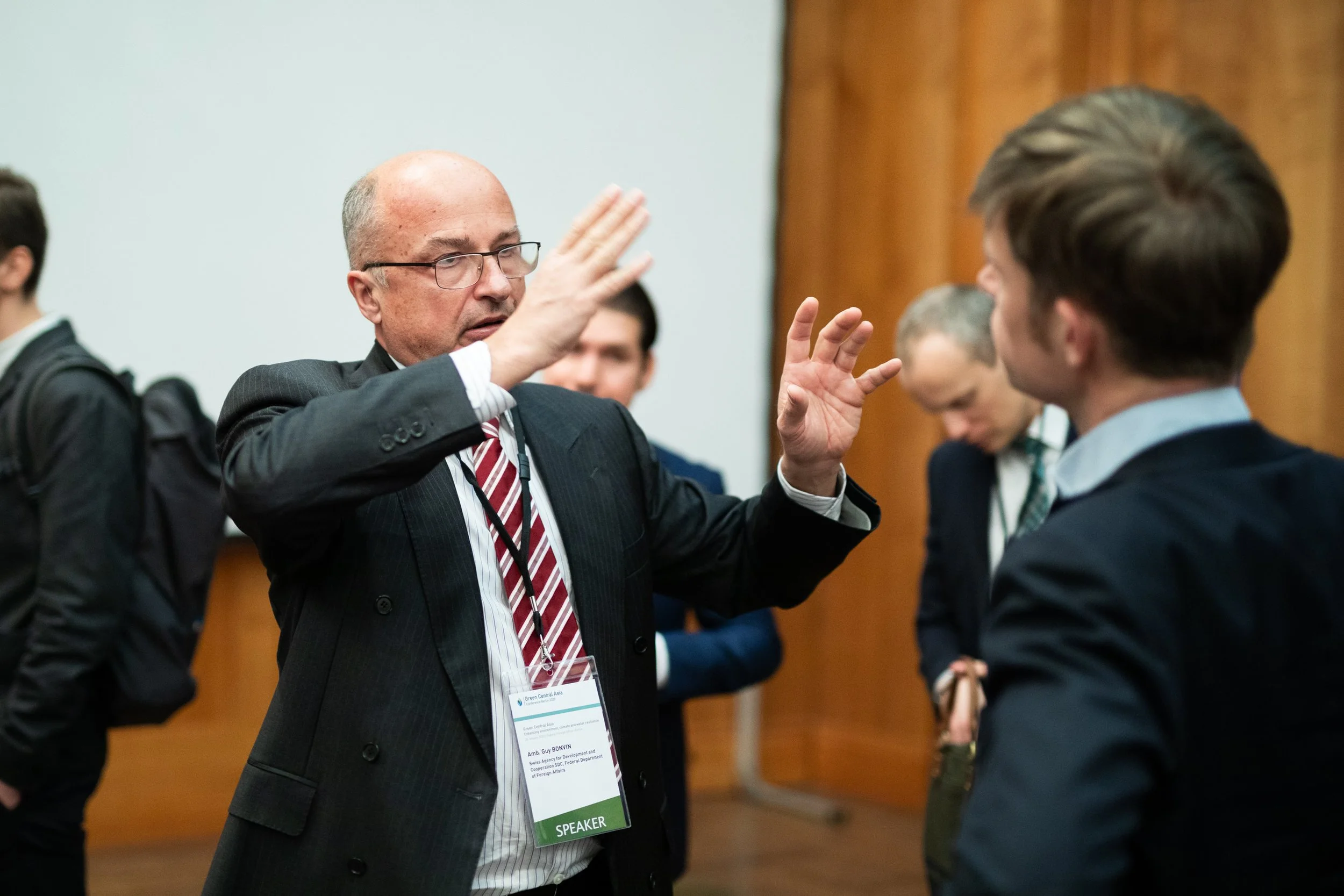 Ein Mann in einem Anzug und mit Namechild spricht mit einem jüngeren Mann bei einer Konferenz. Im Hintergrund sind weitere Menschen, die sich unterhalten.