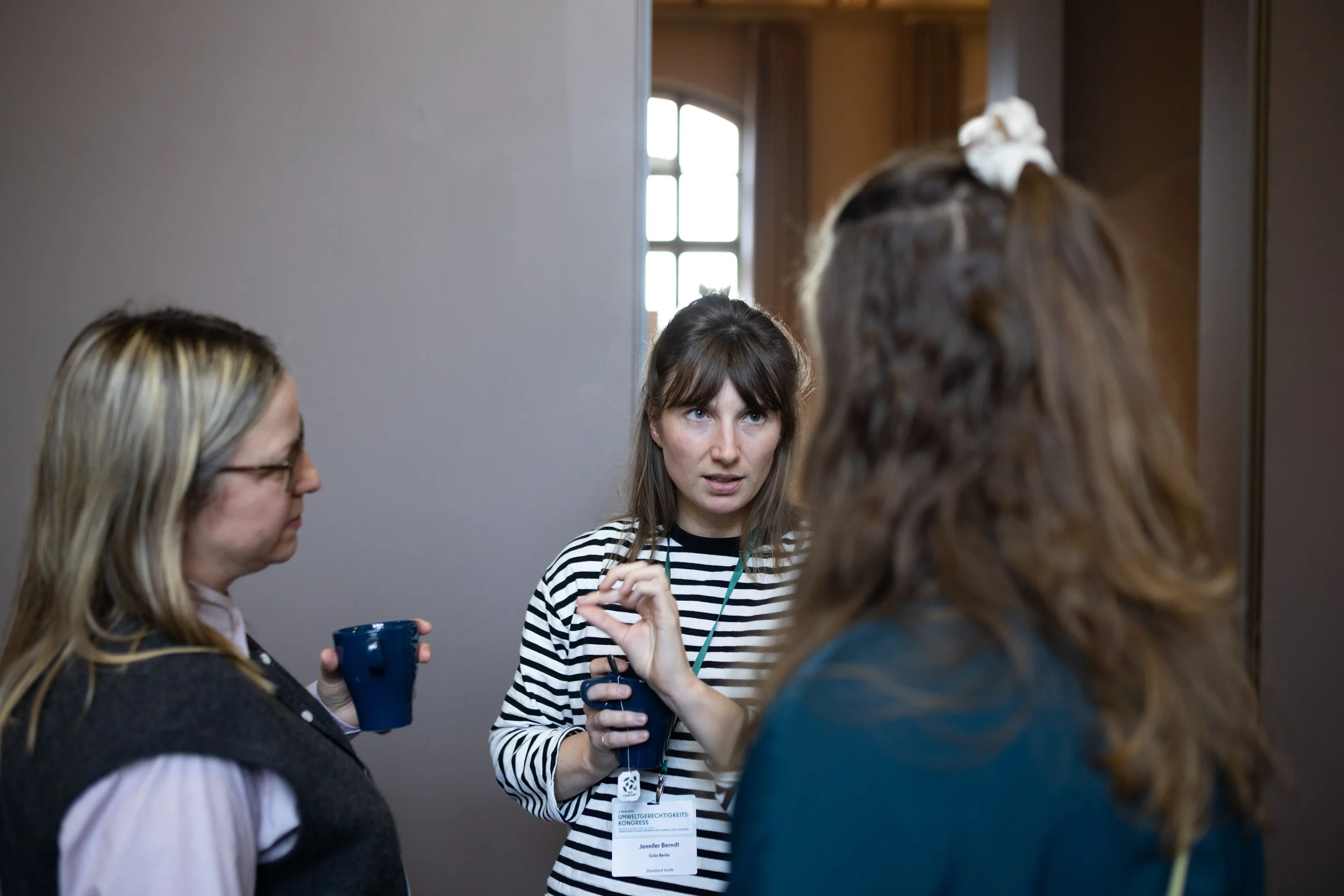 Ein Frauenkongress mit mehreren Frauen im Gespräch, eine trägt ein Namensschild, das sie als Jennifer Berndt identifiziert, die andere hält eine Kaffeetasse.