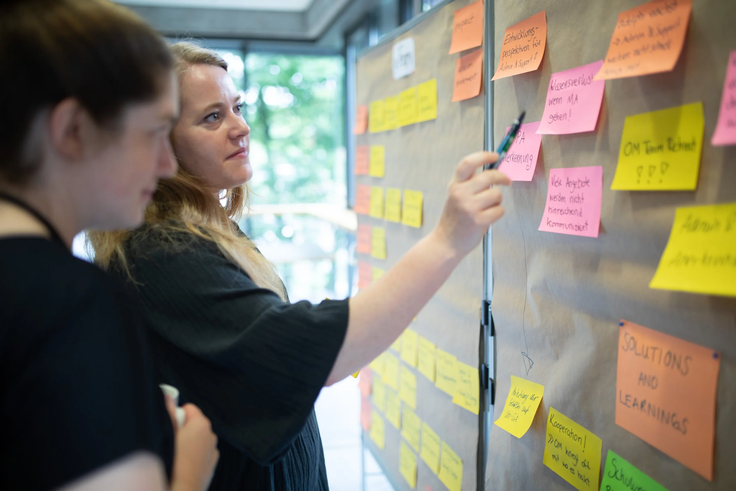 Zwei Frauen schauen auf eine Wand mit bunten Post-its, die verschiedene Notizen und Ideen enthalten.