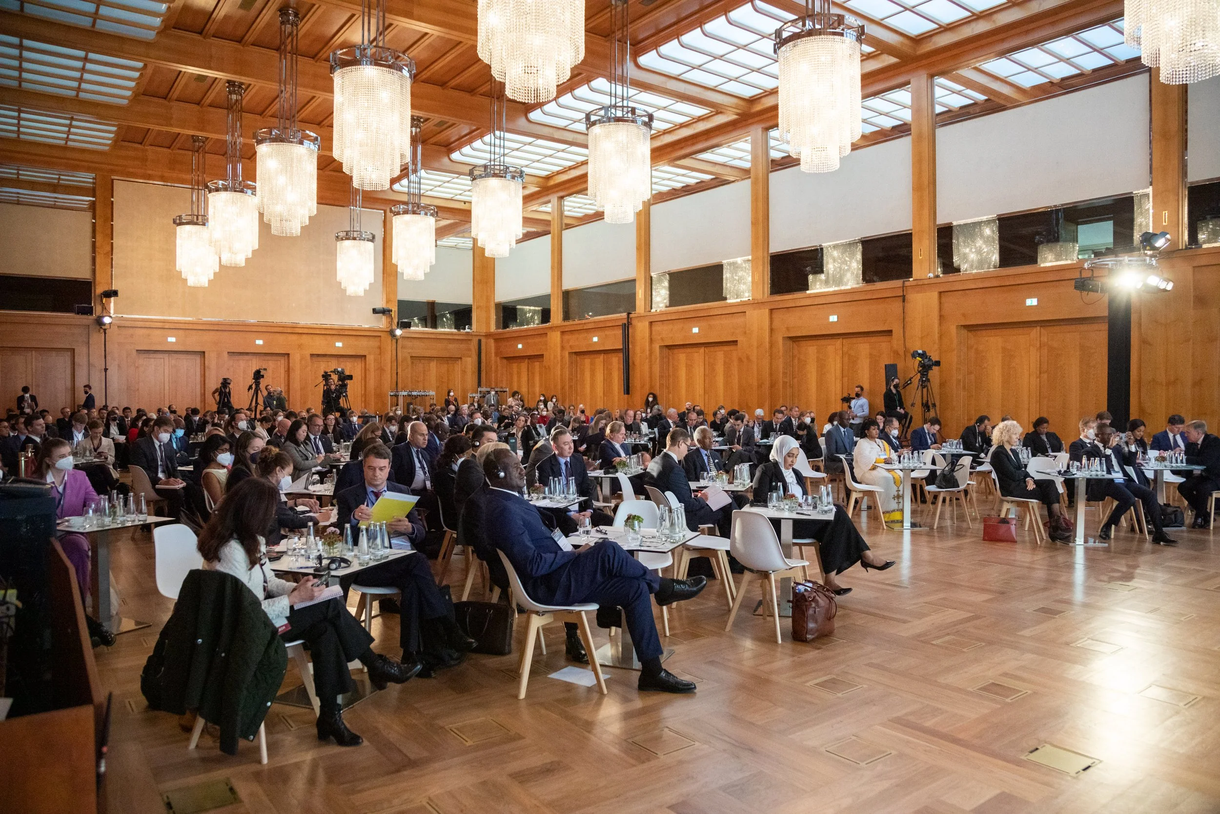 Großer Ballsaal oder Konferenzraum während eines internationalen Treffens mit vielen angereisten Delegierten. Der Raum ist mit Holzwandpaneelen verkleidet, mit mehreren großen Kristallleuchtern und einer Riffeldecke. Viele Menschen sitzen an Tischen,