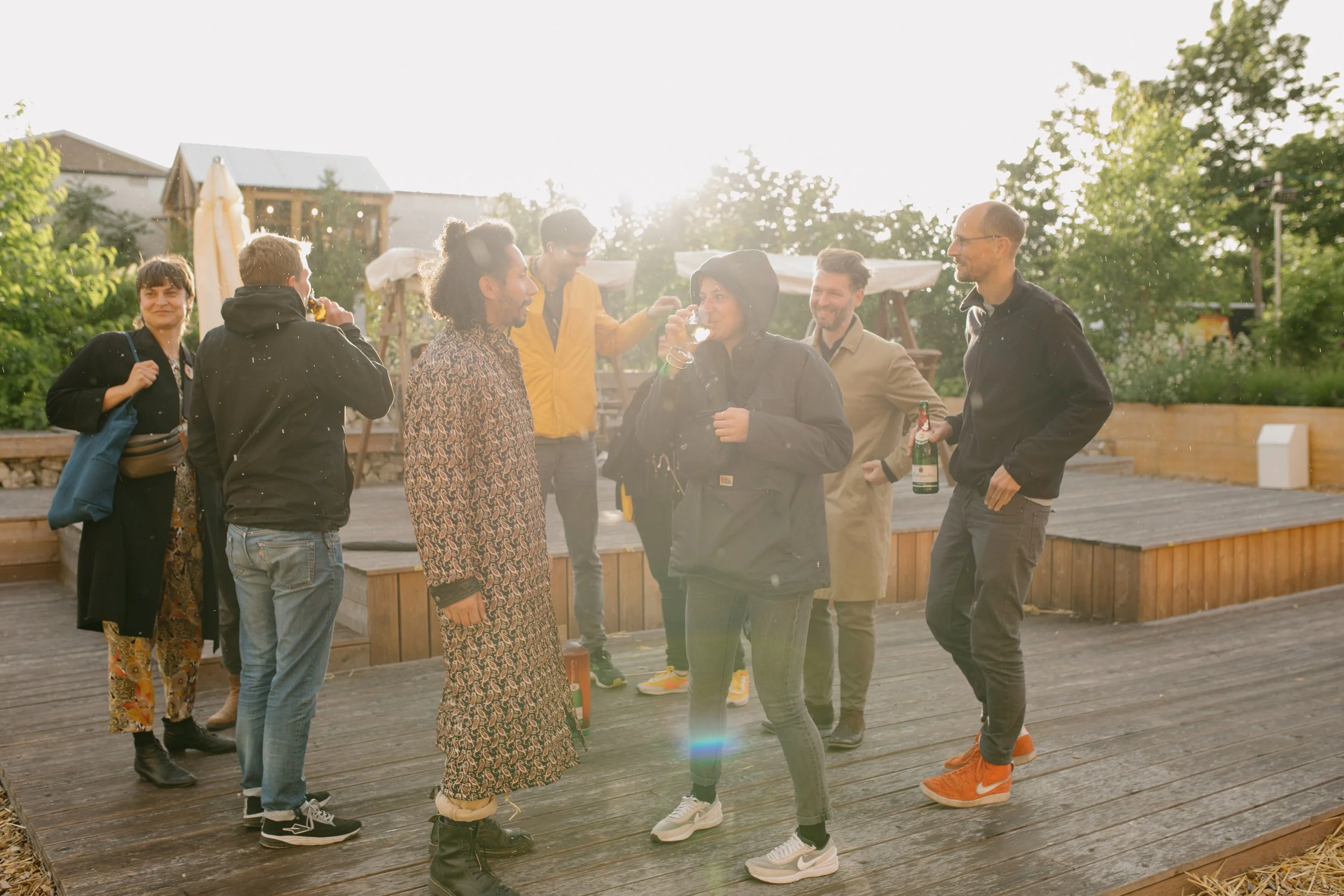 Sieben Menschen unterhalten sich auf einem Holzdeck im Freien bei Sonnenuntergang, umgeben von Bäumen und einem kleinem Spielplatz im Hintergrund.