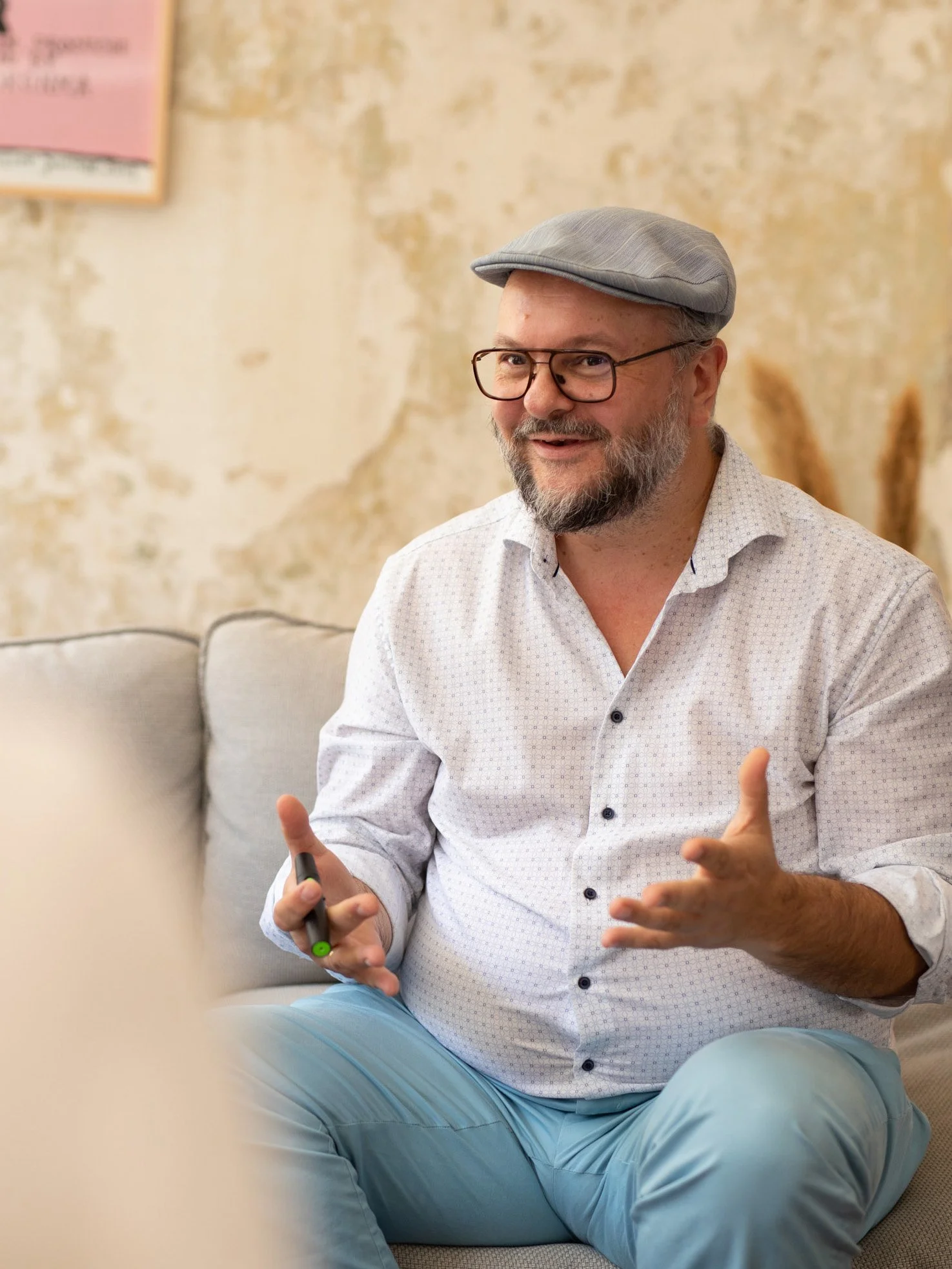 Ein Mann mit Brille und grauem Hut, der sitzend spricht, während er mit einem Marker in der Hand gestikuliert. Im Hintergrund eine Wand mit abblätternder Farbe an einer Couch.