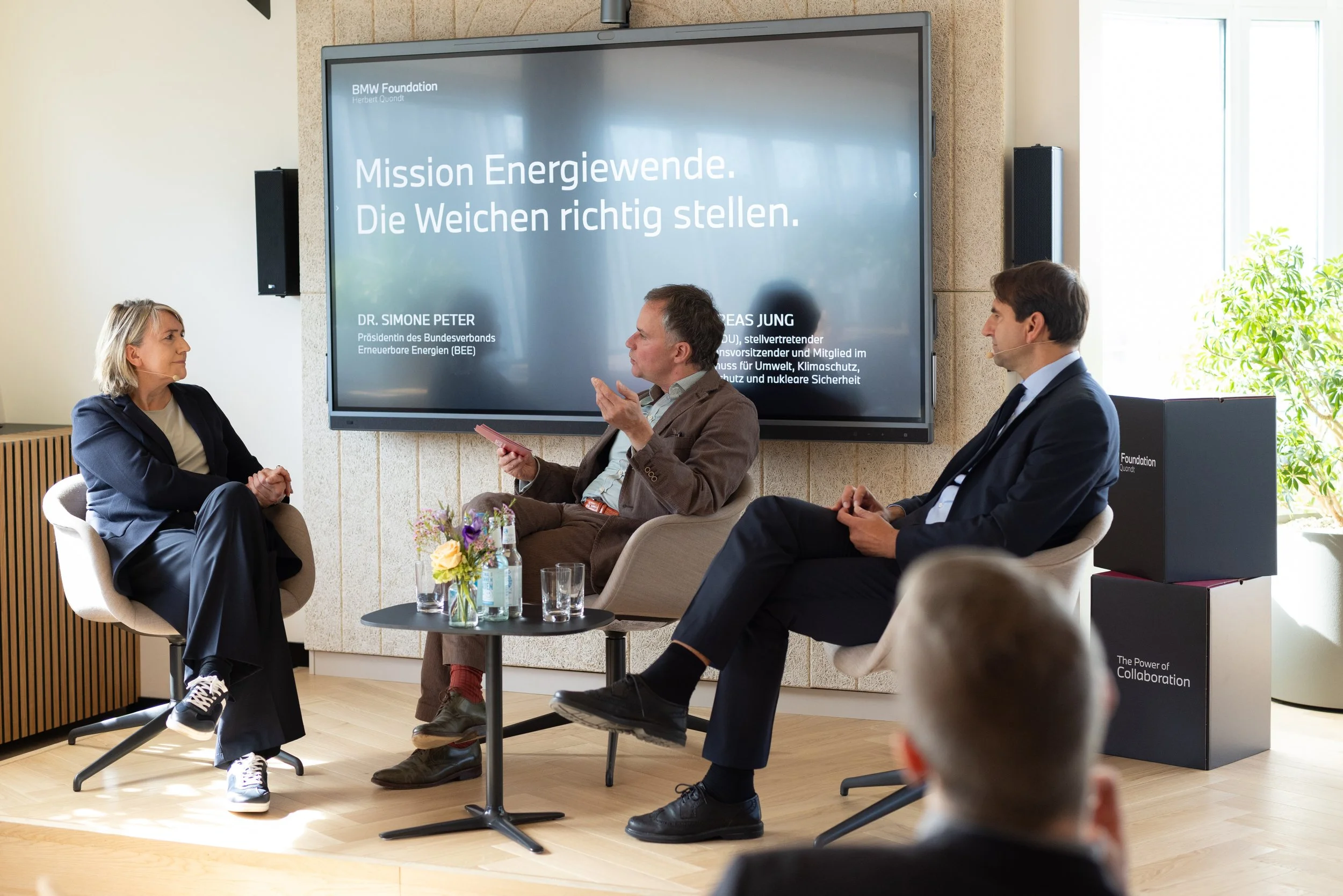 Drei Personen sitzen in einem Gespräch vor einem Bildschirm auf einer Bühne, auf dem steht: 'Mission Energiewende. Die Weichen richtig stellen.' Die Veranstaltung wird von der BMW Foundation Herbert Quandt veranstaltet.